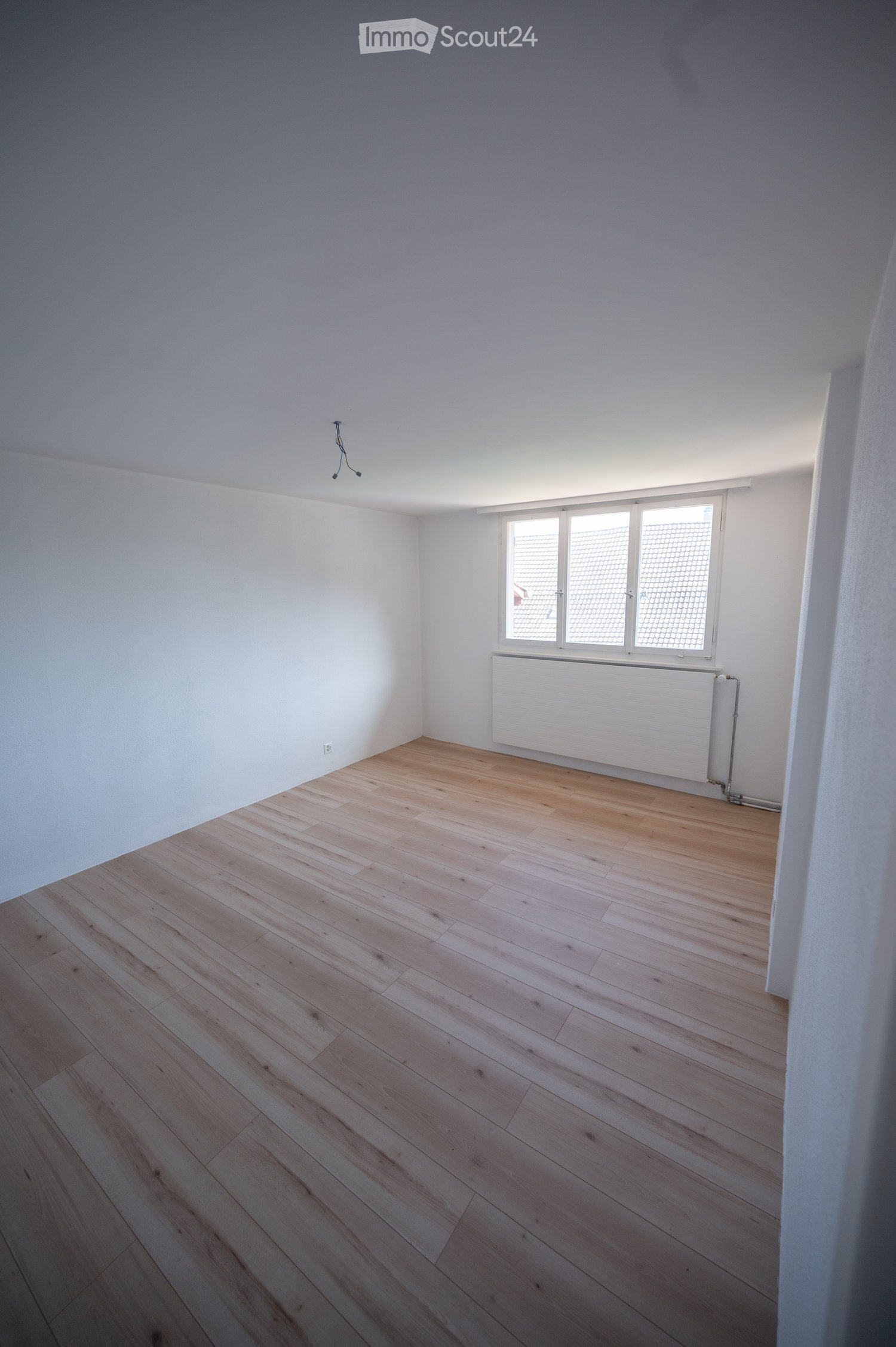 Empty room with light wooden flooring, white walls, and a window with blinds. A radiator is visible below the window.