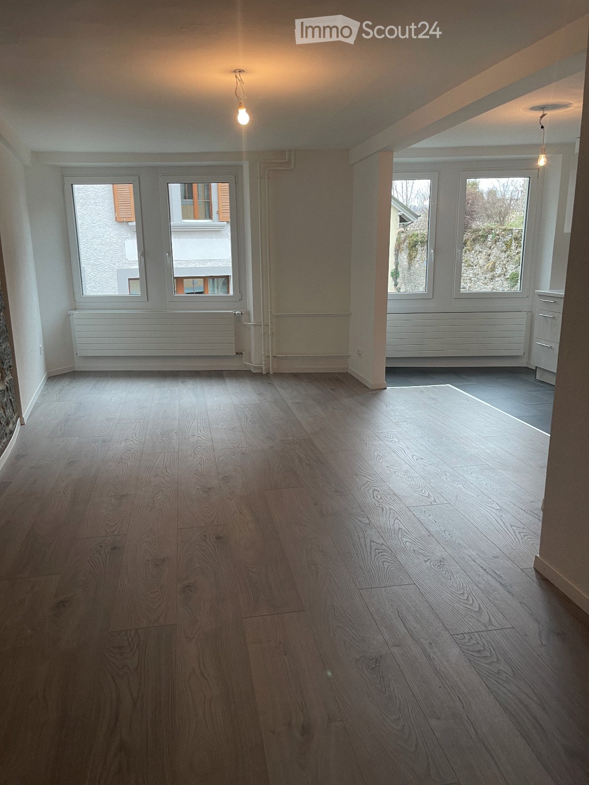 Empty room with wooden flooring, white walls, large windows, radiators, and exposed pipes.