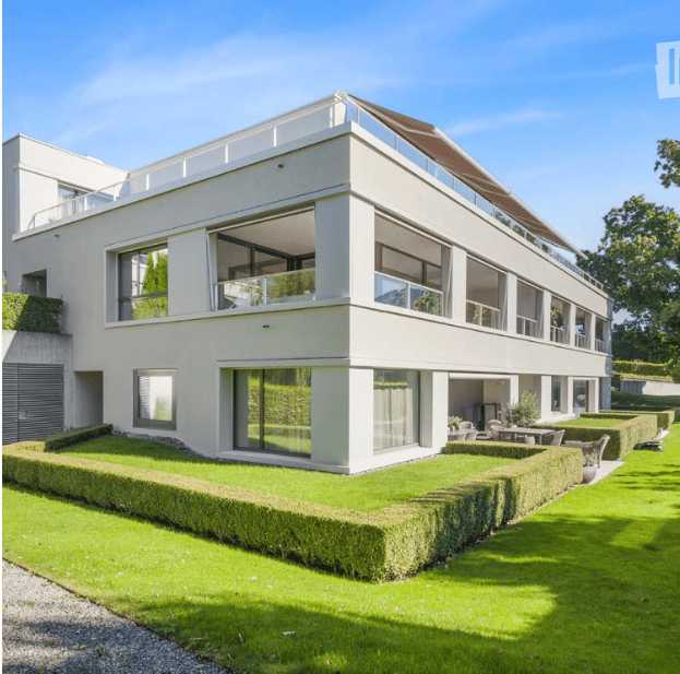 Modern two-story house with white exterior, large windows, and a flat roof. The house features a well-maintained lawn and neatly trimmed hedges in the front yard.