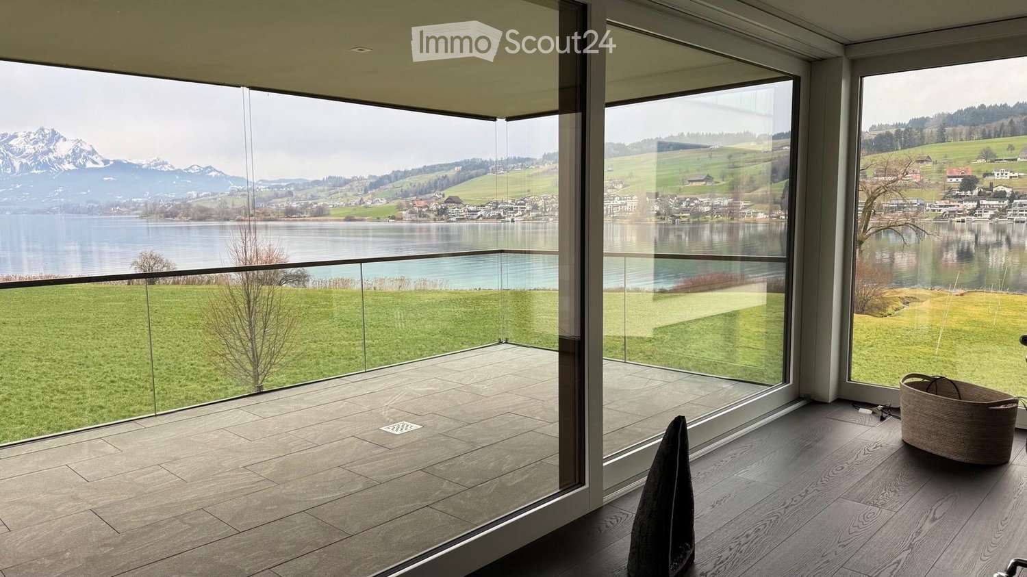 Modern balcony with large glass walls offering scenic views of a lake and mountains, featuring a wicker basket and a folded garment on the floor.