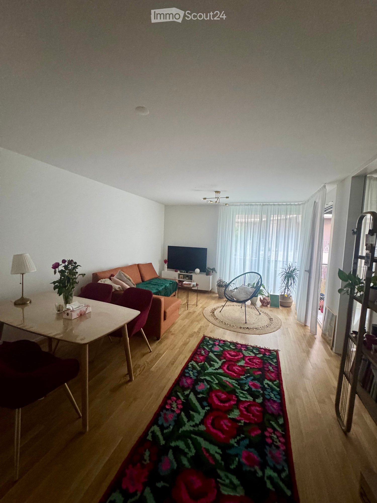A modern living room with a wooden floor, a sofa, a dining table, a lamp, a television, and decorative rugs.