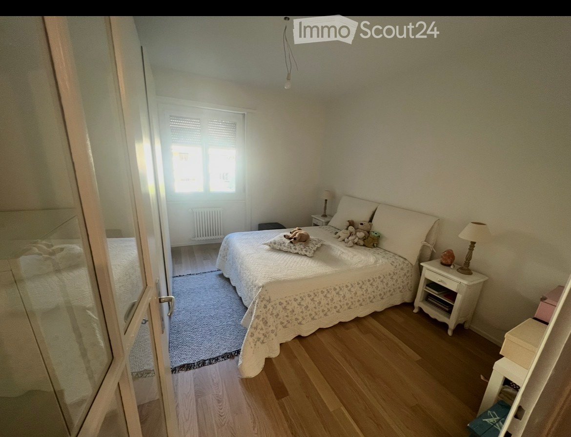 Cozy bedroom with wooden flooring, a double bed with white bedding and stuffed toys, a nightstand with a lamp, and a radiator beneath a window.