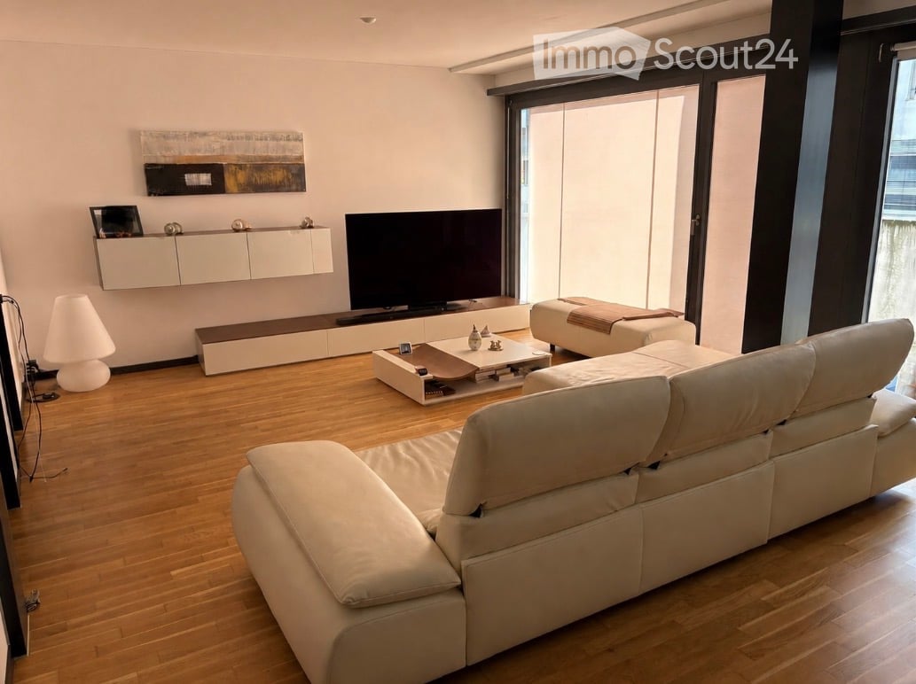 Modern living room with light-colored furniture, including a large sectional sofa, coffee table, ottoman, and a TV stand. The room features a sleek wall-mounted cabinet and a contemporary floor lamp.