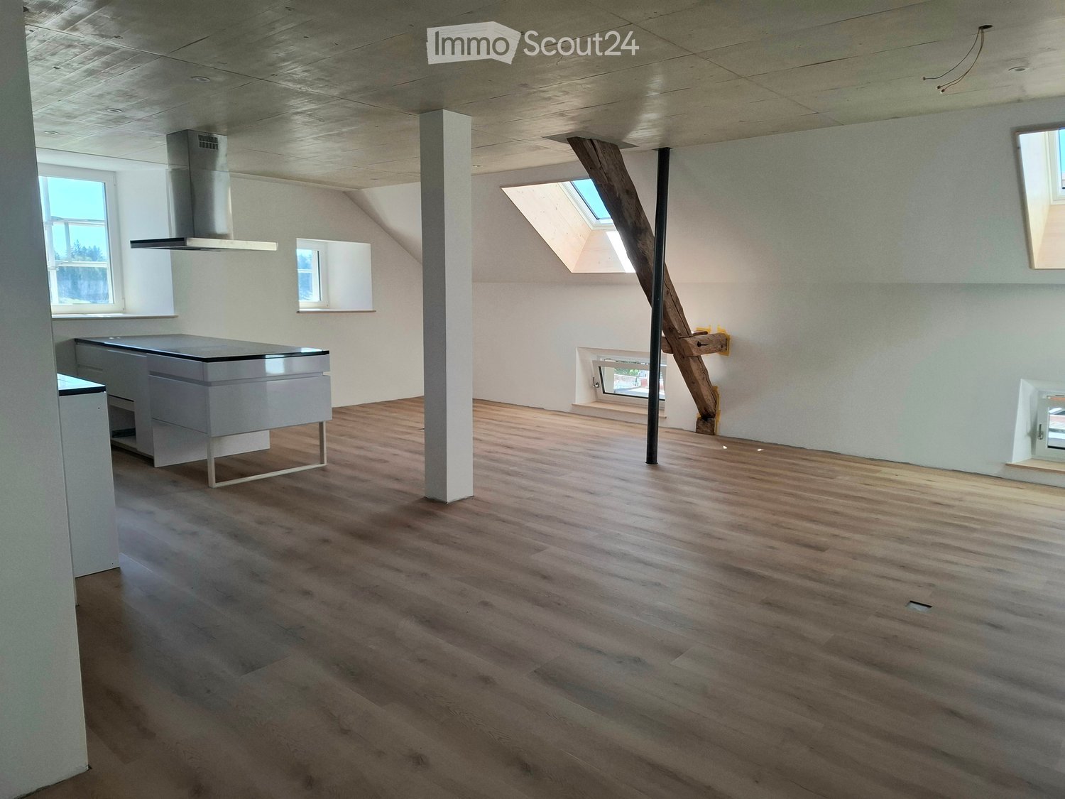 Open-plan space with wooden flooring, white walls, and modern kitchen units. Features include a large window, skylight, and a central support column.