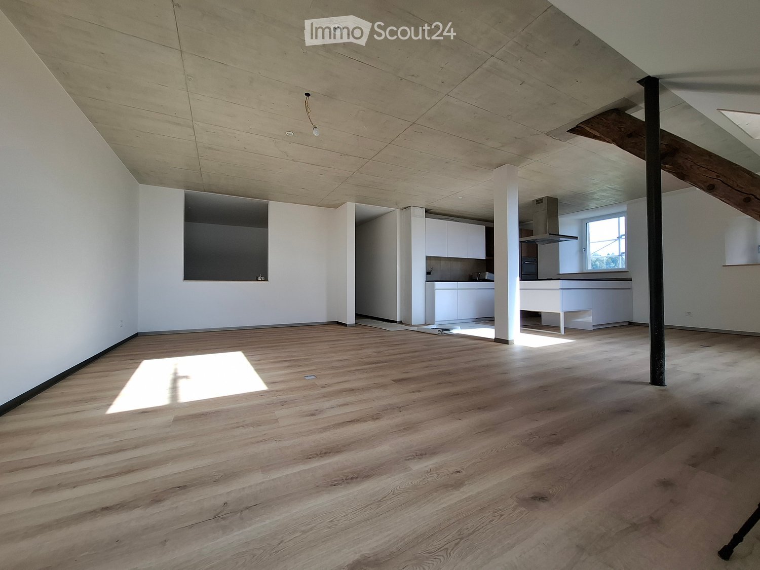 Open, unfurnished room with wooden flooring, white walls, and exposed wooden beams on the ceiling. The room features a large window allowing natural light to enter, and a small kitchen area is visible in the background.