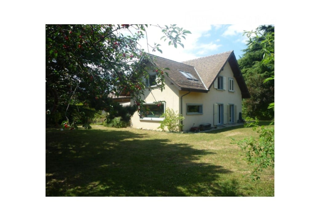 Single-story house with a pitched roof, surrounded by a well-maintained lawn and trees. The house features multiple windows and a small patio area.