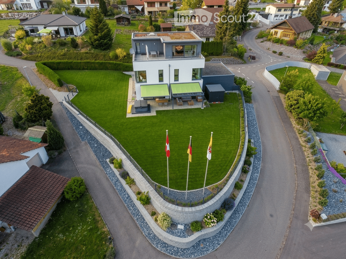 Aerial view of a modern house with a large, well-maintained lawn, surrounded by a stone wall and flags, with neighboring houses and a winding road visible.