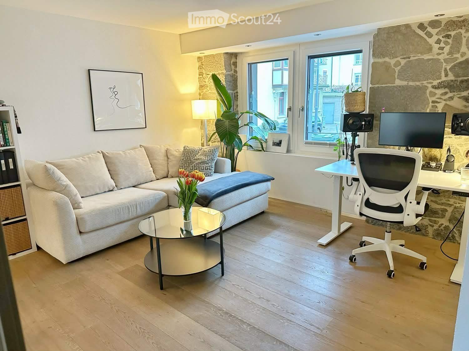 Modern living room with a white sectional sofa, glass coffee table, white desk with ergonomic chair, large windows, and a stone accent wall.