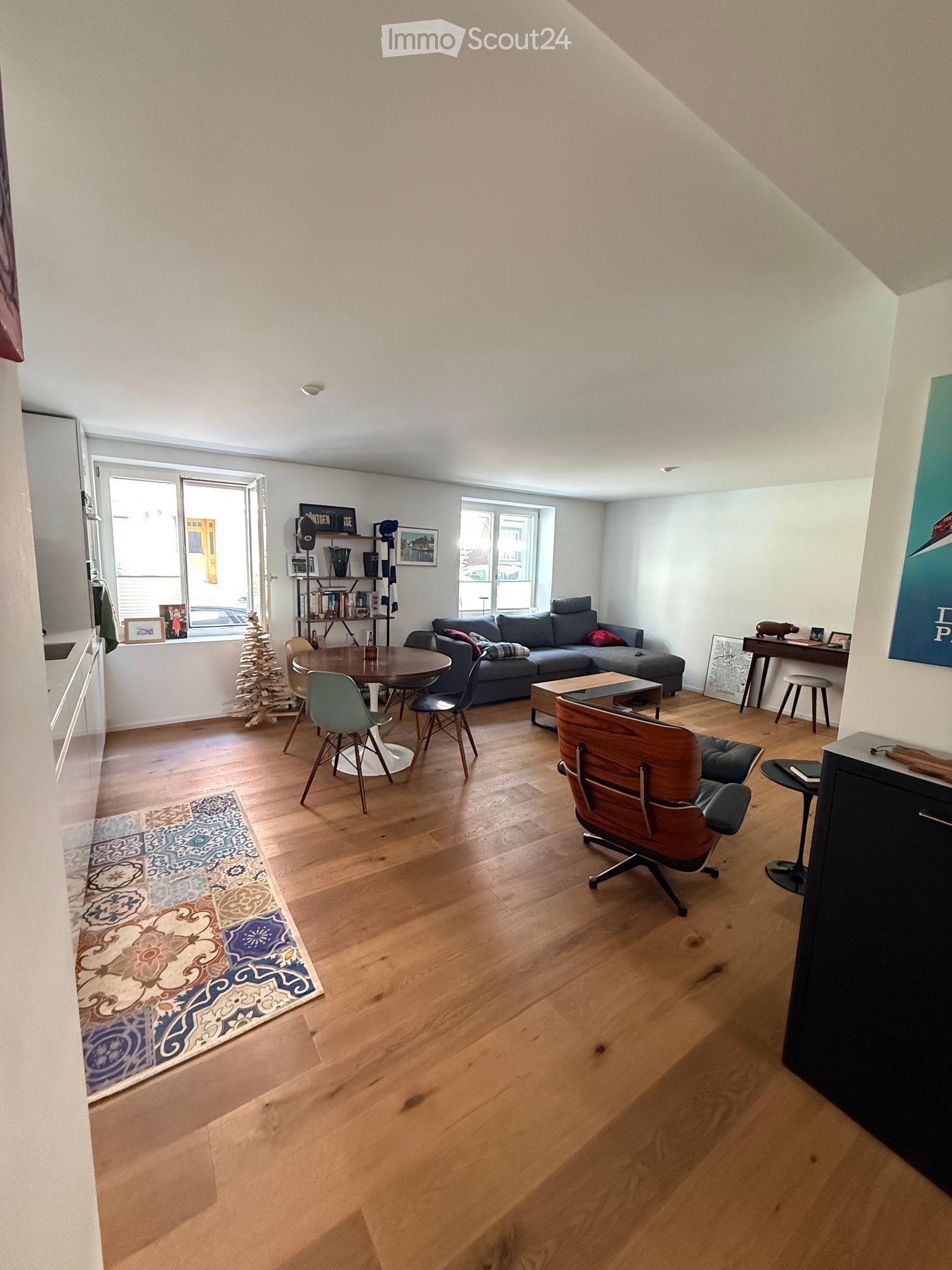 Bright living room with wooden flooring, large windows, a grey sectional sofa, a round dining table with chairs, a wooden armchair, a small shelf with books and decor, and a patterned rug.