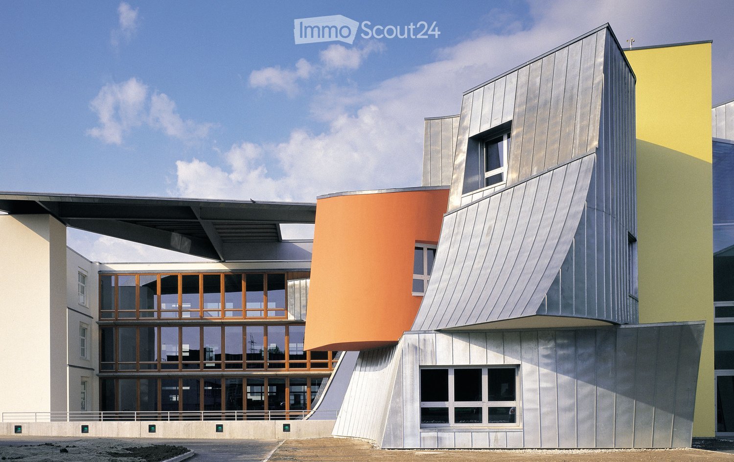 Modern, multi-story building with a combination of metallic, orange, and green facades, large windows, and a raised ground floor entrance.