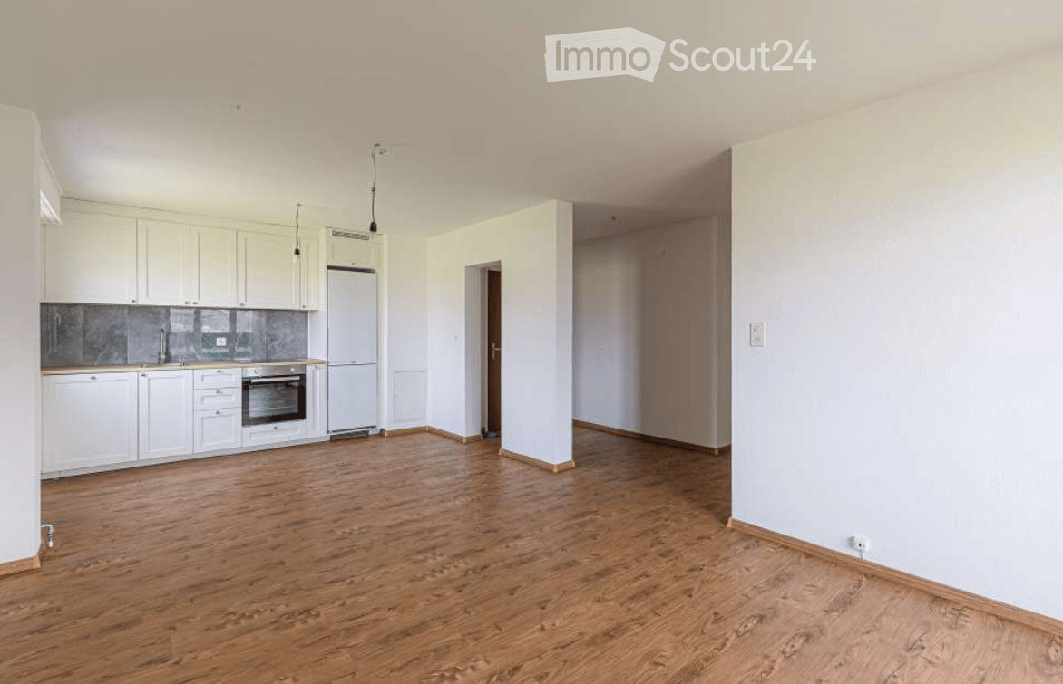 Empty room with wooden flooring, white walls, and a small kitchen area featuring white cabinets, a sink, and a refrigerator.
