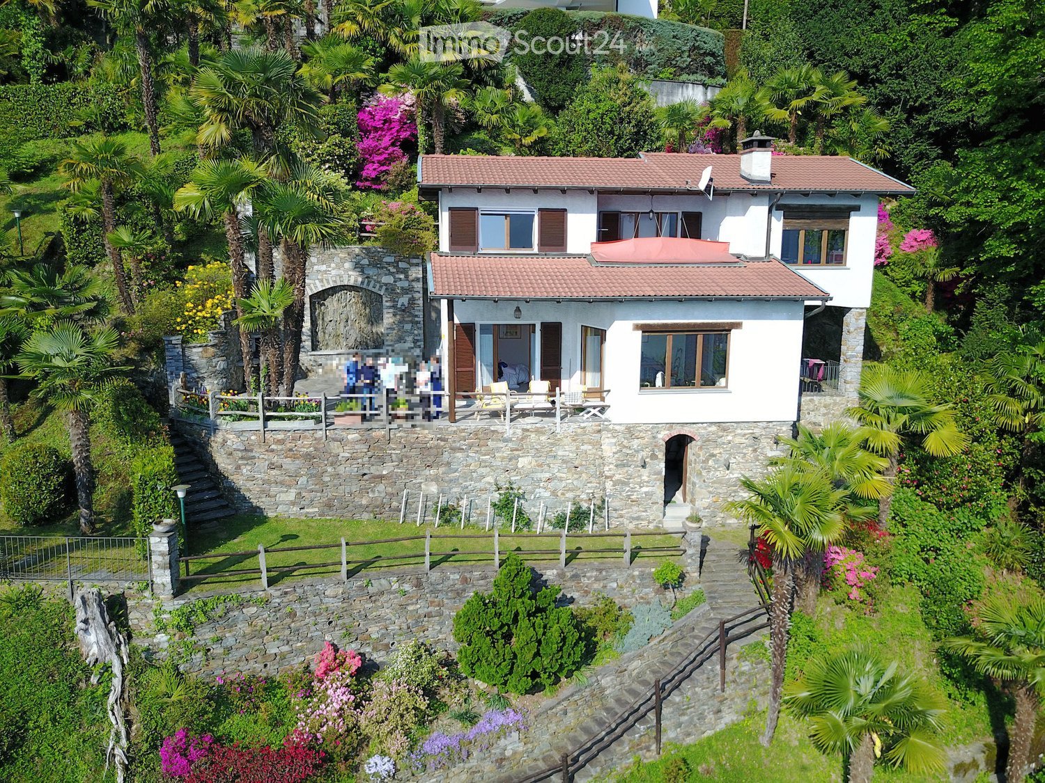2 story white house with brown roof, surrounded by garden with flowerbeds, stone wall, wooden fence, and trees, terrace with chairs, located on a hillside