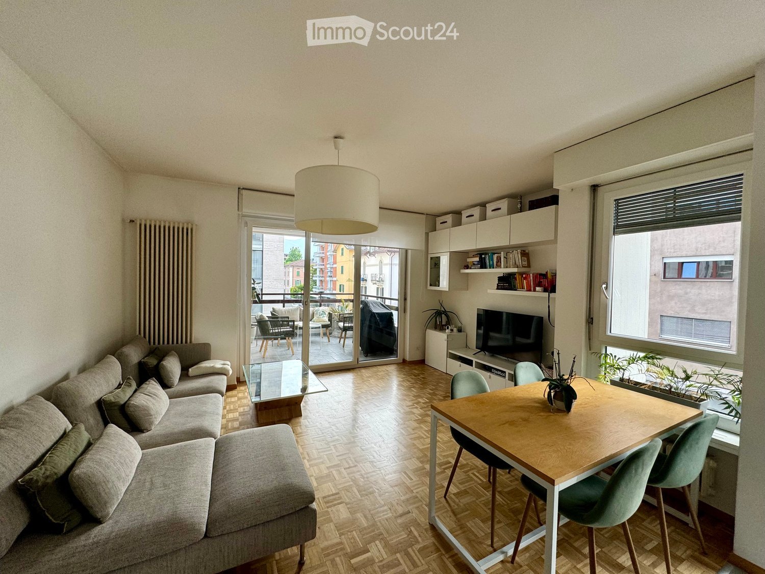 Living room with a grey sectional sofa, glass coffee table, wooden dining table with chairs, TV, bookshelf, and a balcony with outdoor furniture visible through the sliding glass door.