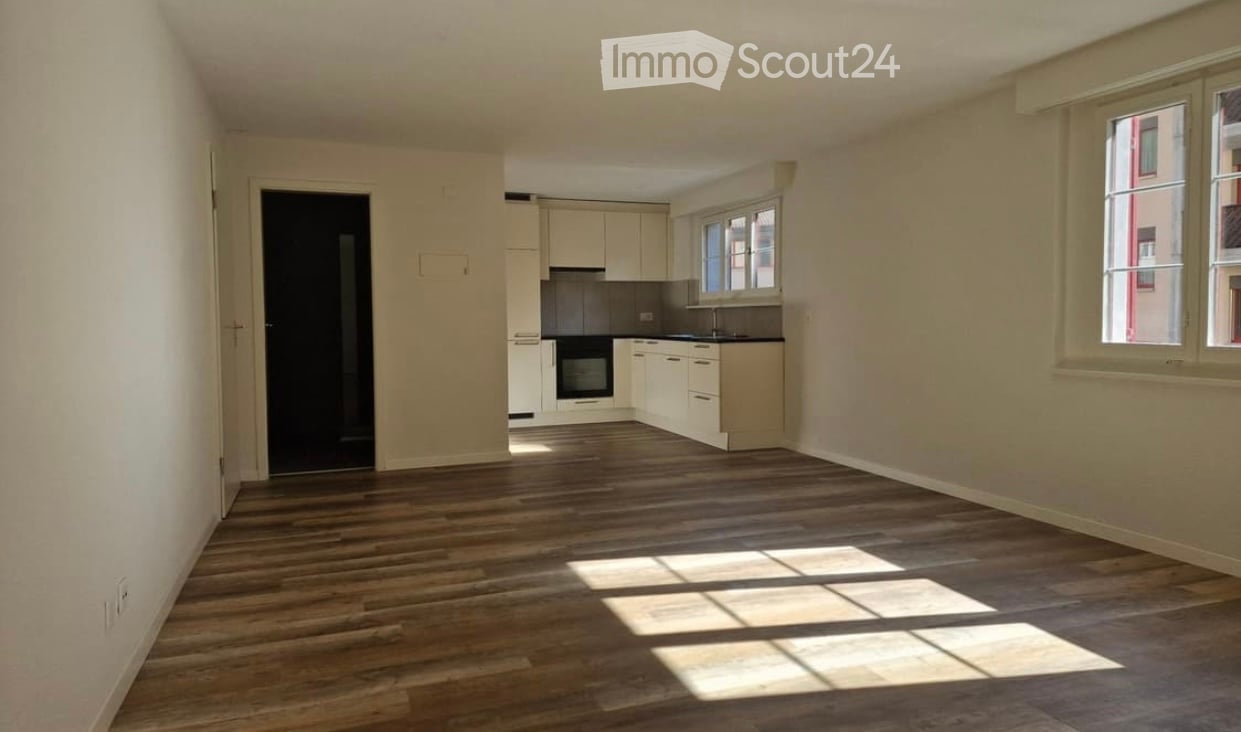 Bright, open-plan room with wooden flooring, white kitchen cabinets, and a built-in oven. Large windows allow natural light to fill the space.