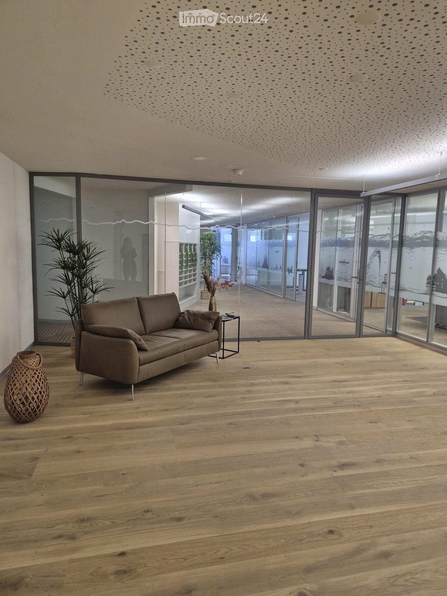 Modern living room with light wooden flooring, large glass sliding doors, a brown sofa, side table, decorative vase, and plants.