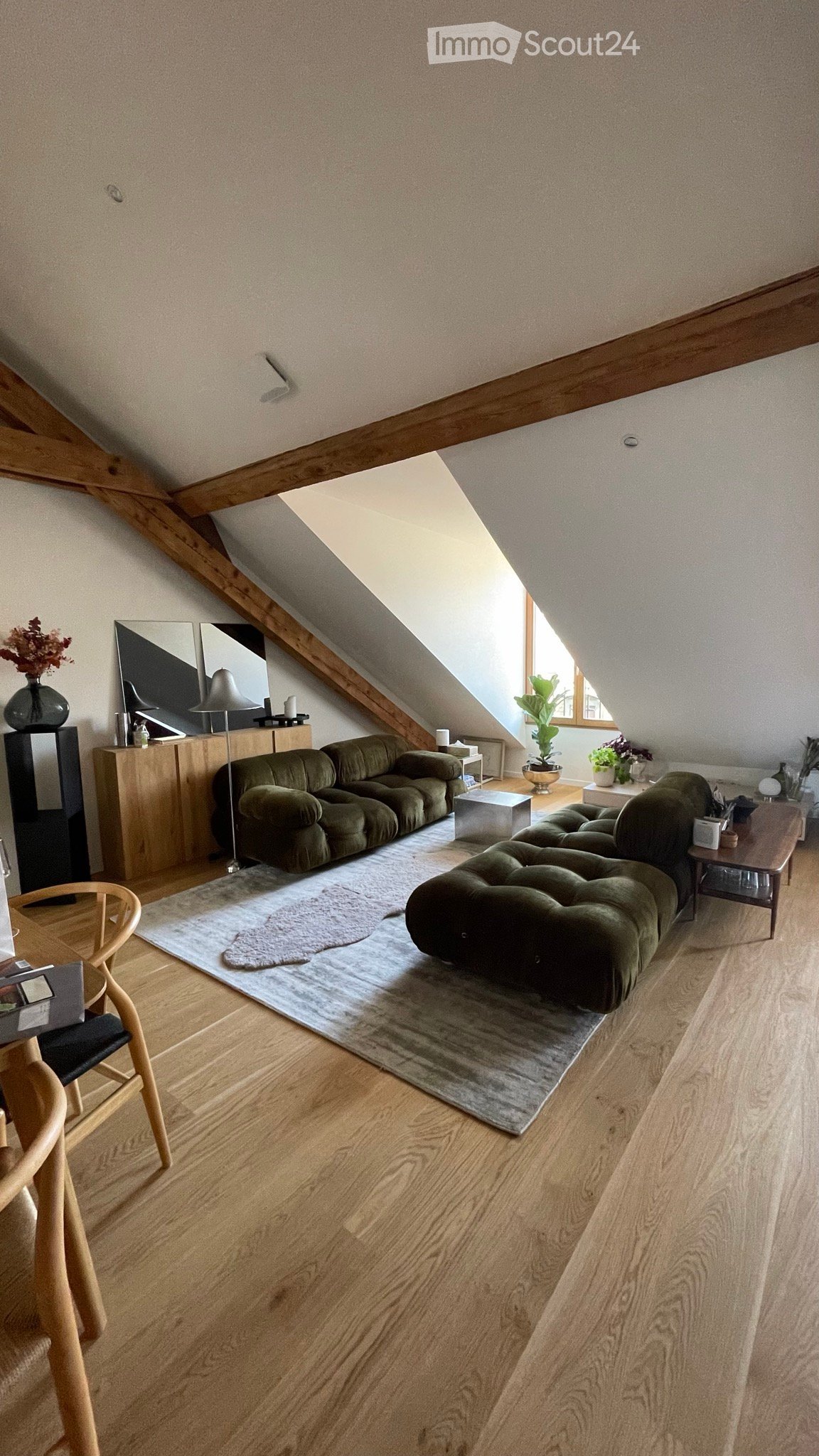 Living room with wooden floors, slanted ceiling, large window, green sofa, side table, lamp, and decorative items.