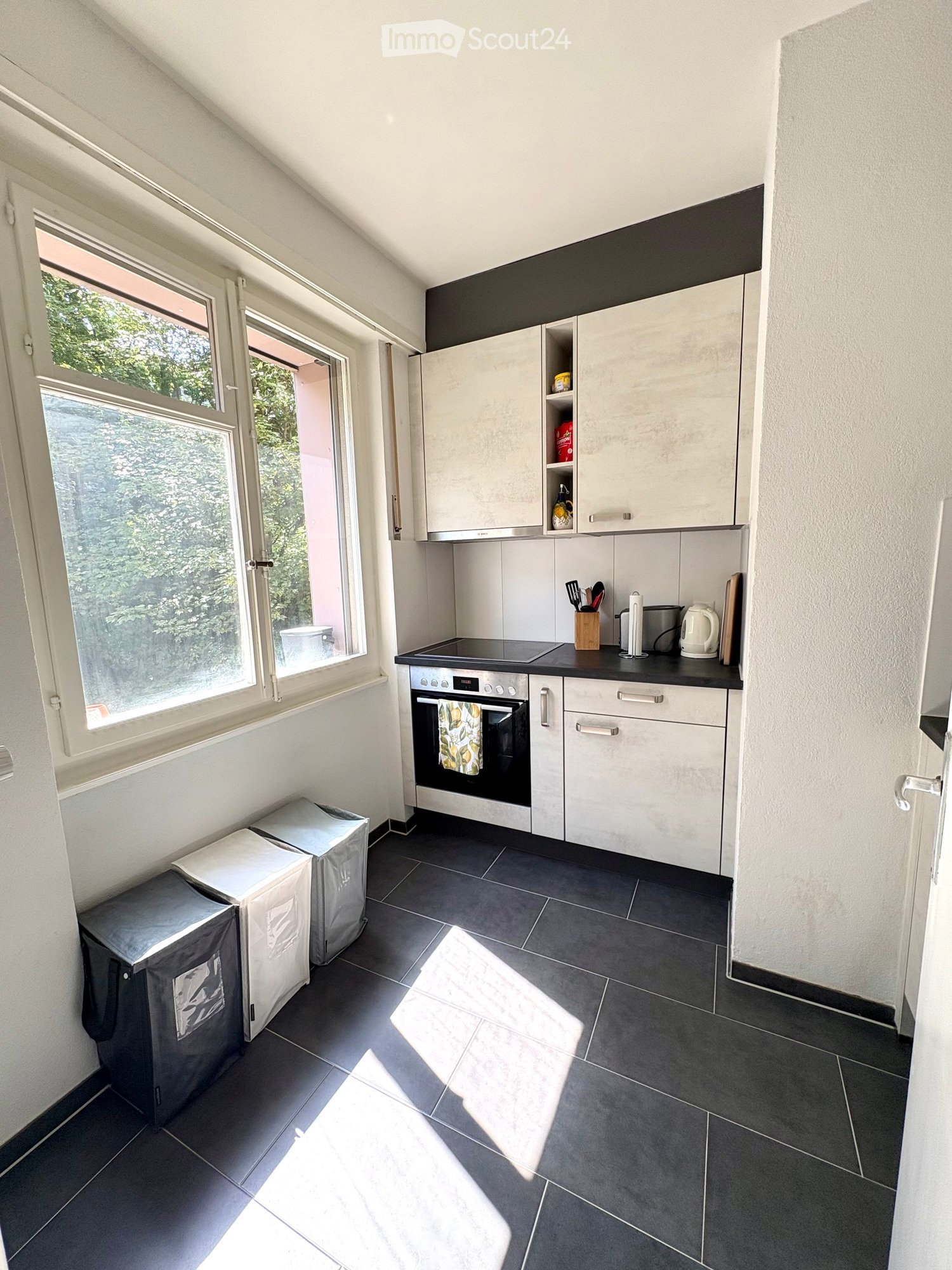 Modern kitchen with white cabinets, black countertop, and stainless steel appliances. Features an oven, microwave, and various kitchen utensils on the counter. The room has large windows allowing natural light to fill the space.