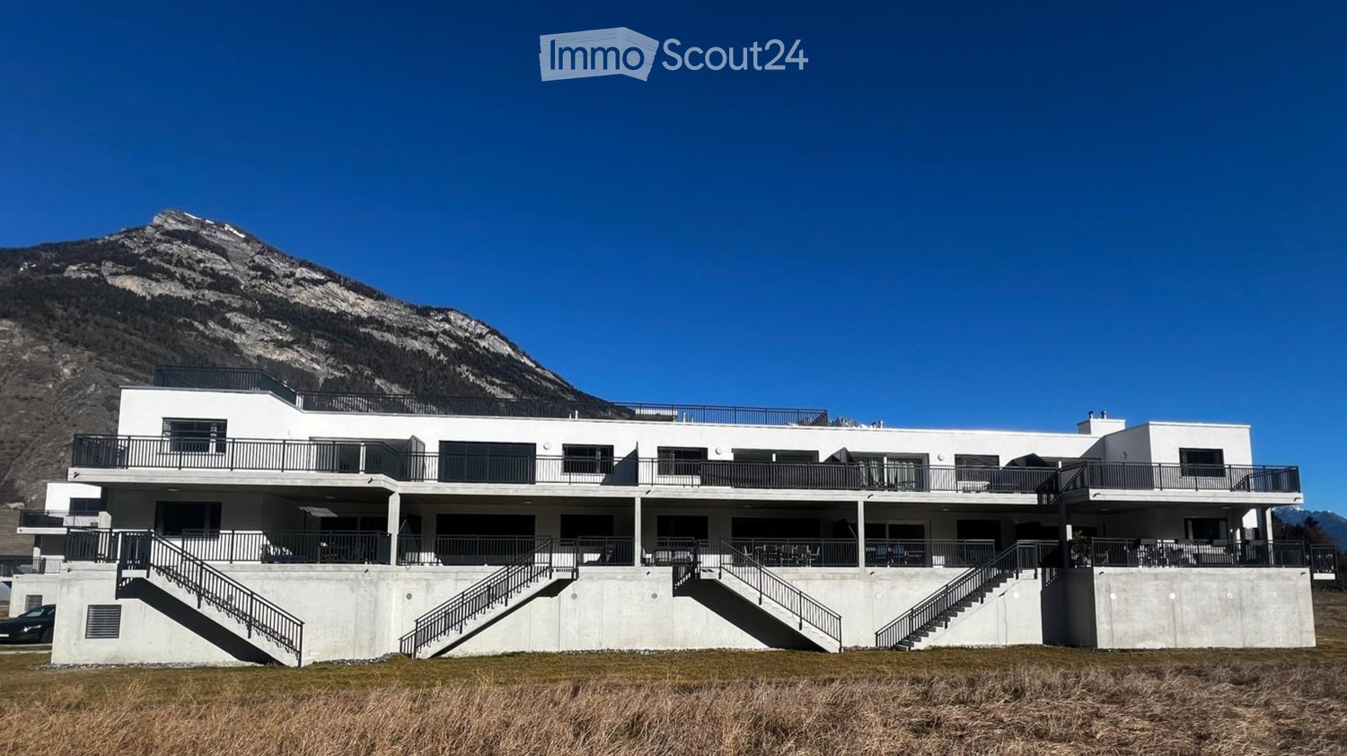 Modern two-story building with white exterior, multiple balconies, and a scenic mountain view in the background.