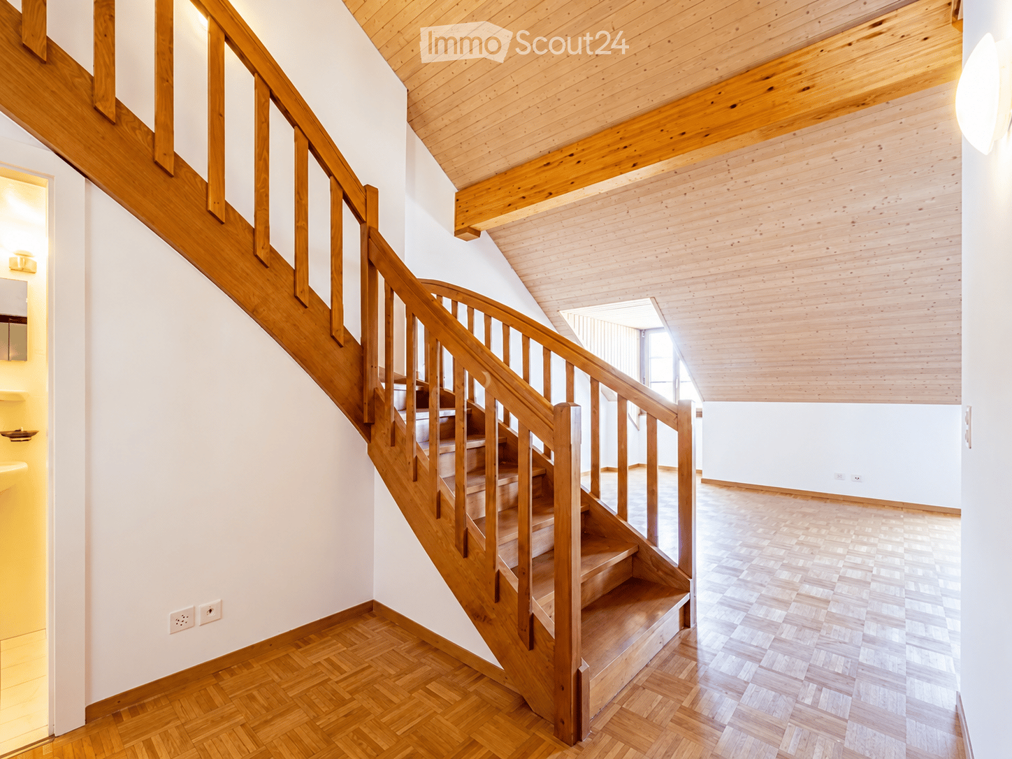 Spacious hallway with wooden staircase, light wooden ceiling, and polished wooden flooring. The staircase features a wooden handrail and steps, leading upwards. The walls are painted white, and the area is well-lit with natural light coming from a window