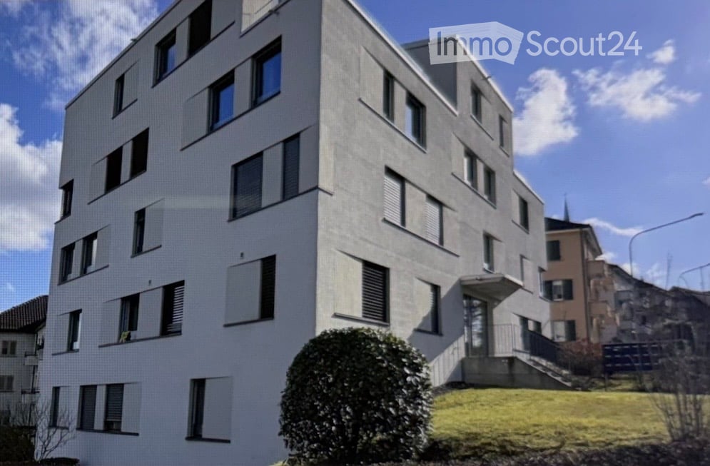 Modern multi-story residential building with white facade, numerous windows, and a small landscaped area in front.