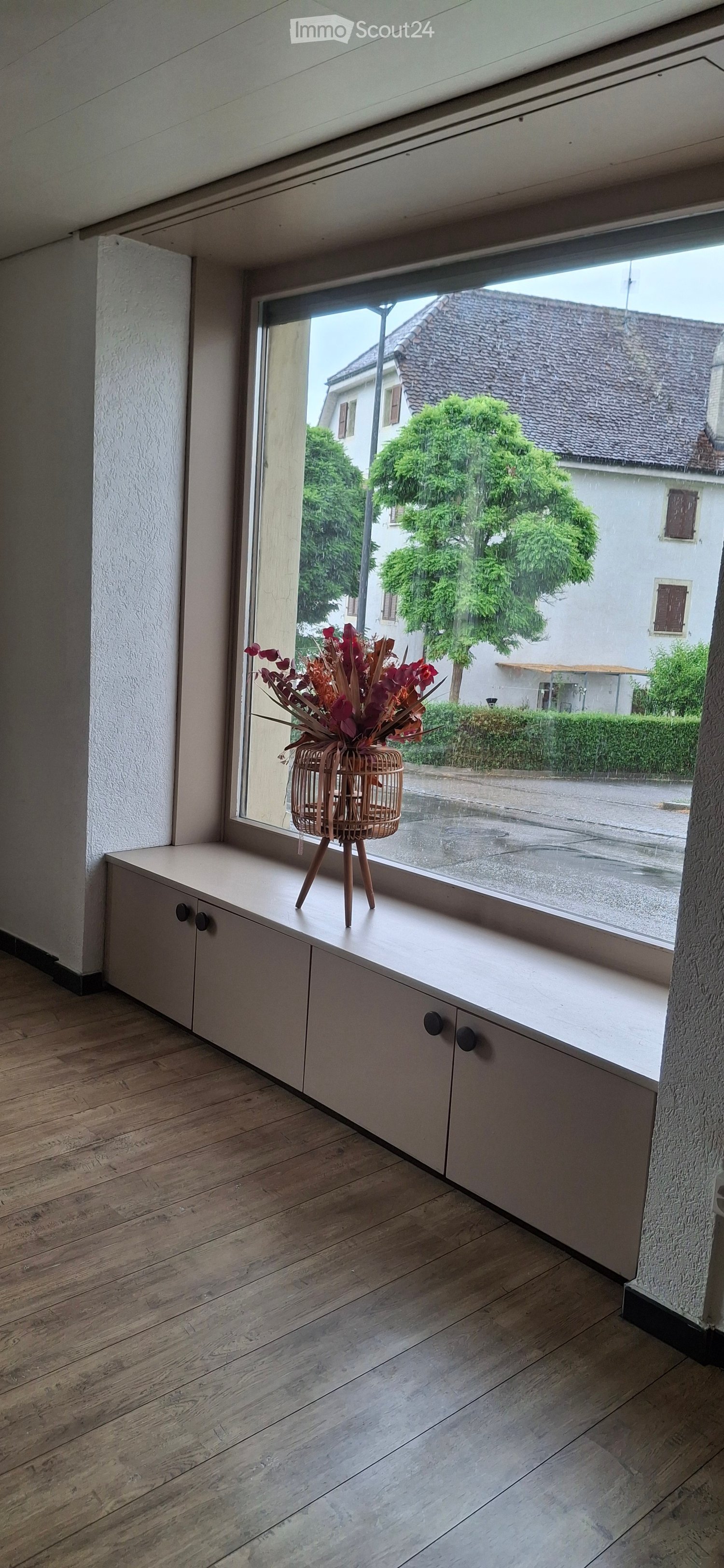 A modern, minimalist room with light wooden flooring and white cabinetry along a large window. A decorative flower arrangement in a woven basket sits on the windowsill.
