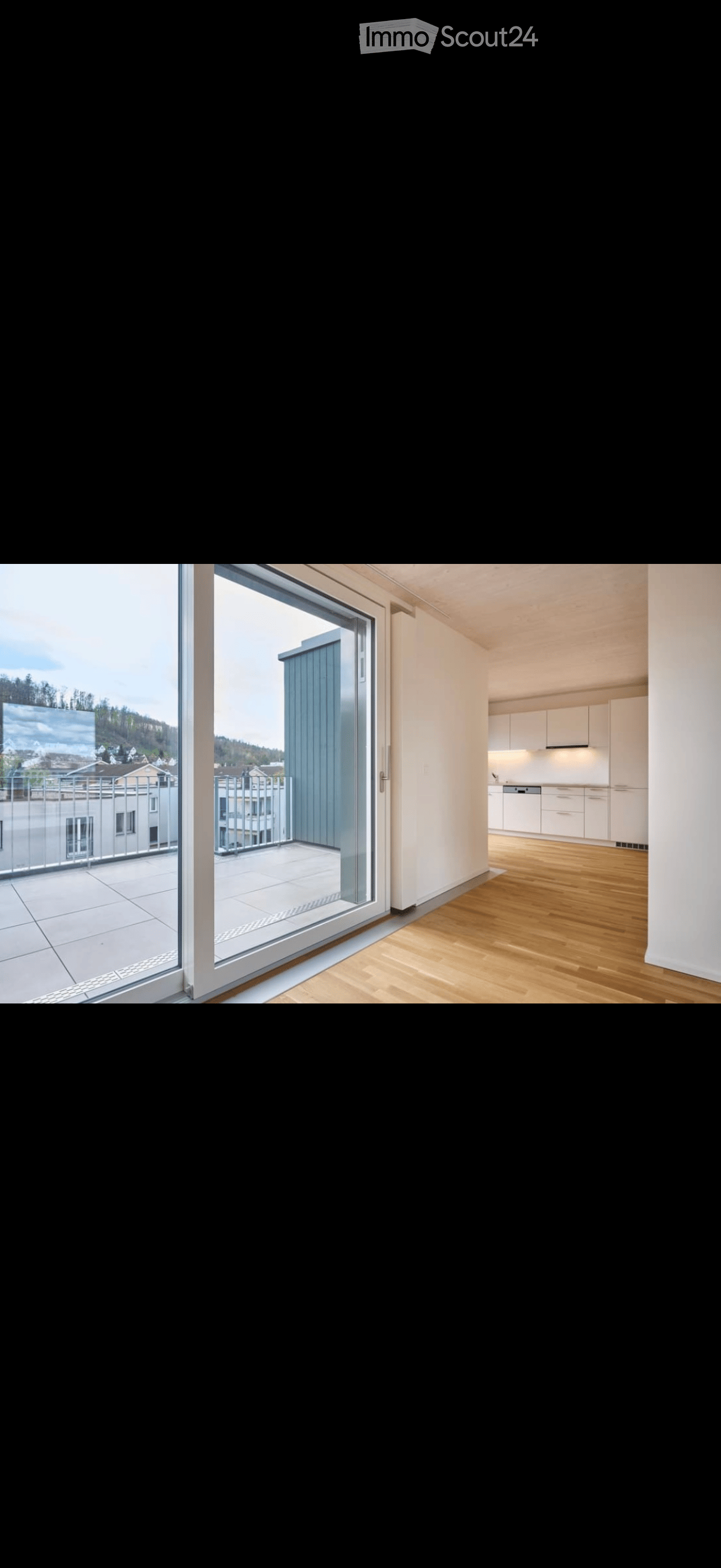 Modern interior with wooden flooring, large sliding glass door leading to a balcony, and a small kitchen area with white cabinets.