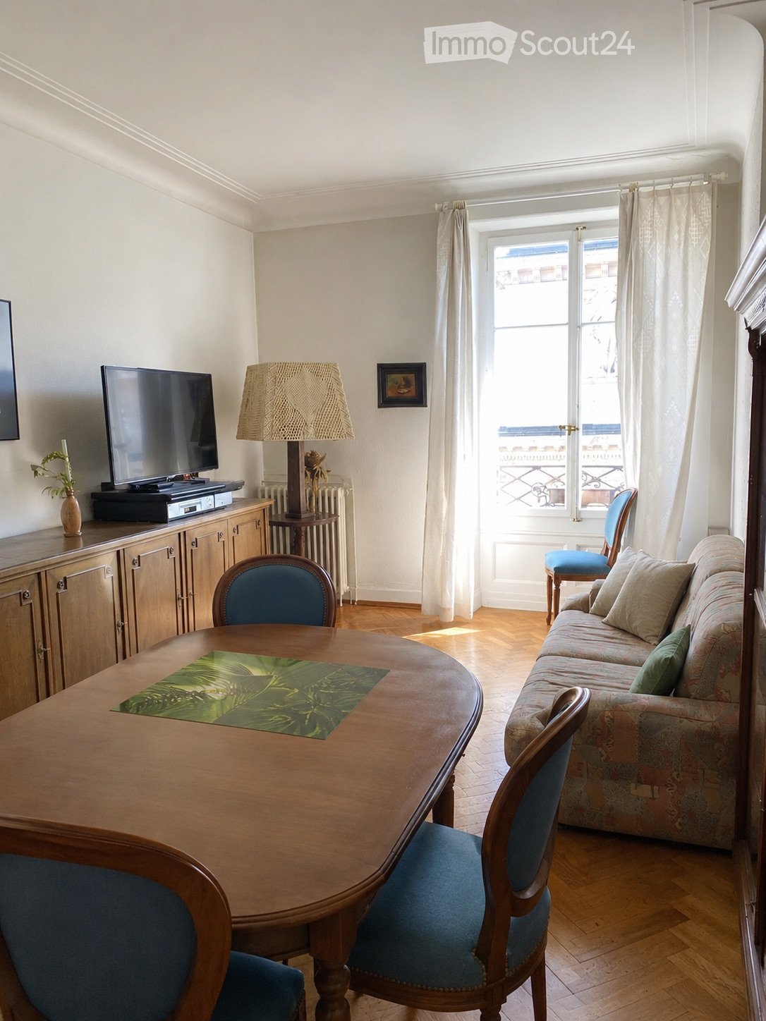 A well-lit living room with wooden flooring, a patterned sofa, a round wooden table with chairs, a lamp, a television on a cabinet, and a vase with flowers near the window.
