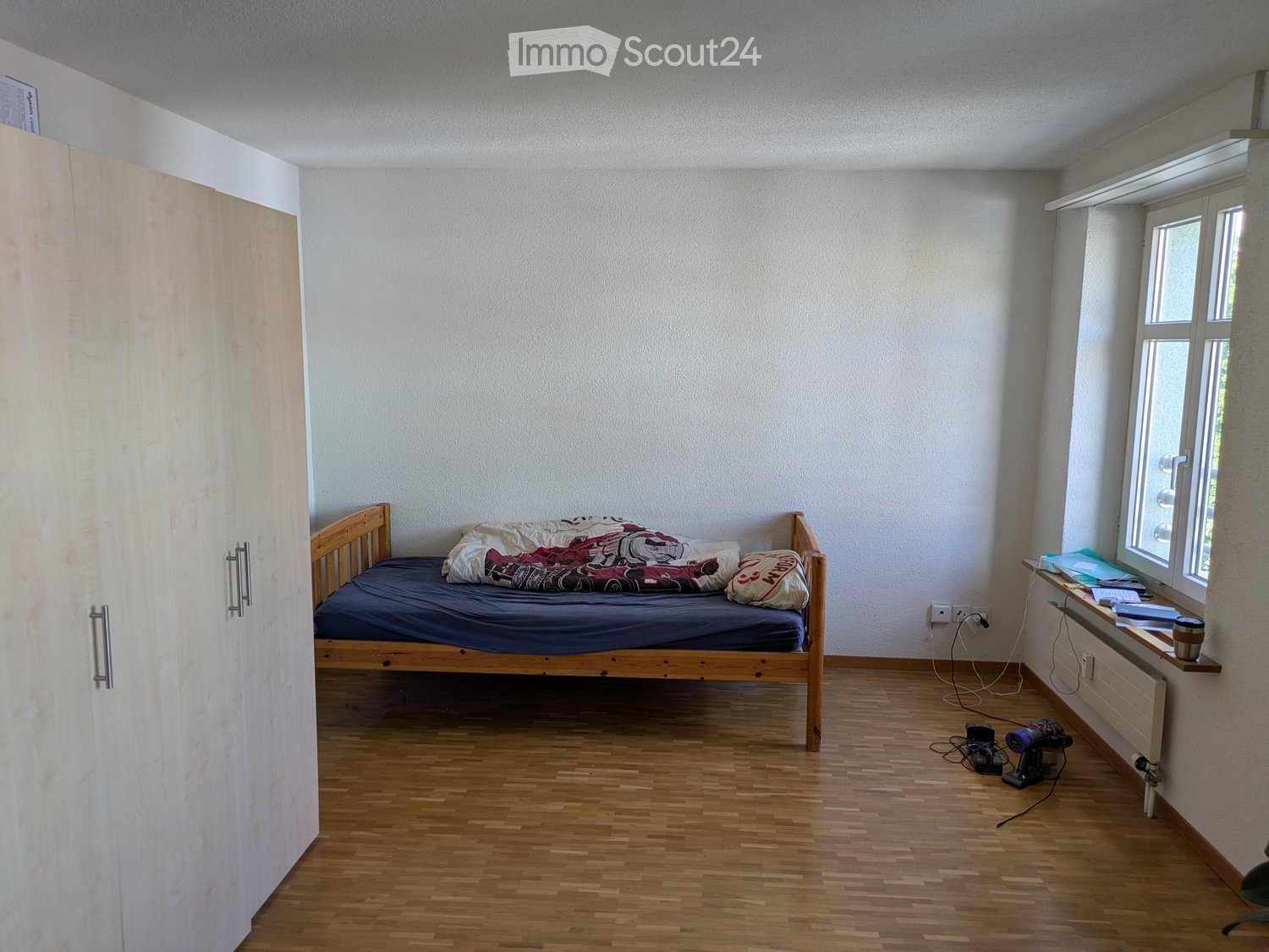 A small bedroom with a wooden bed, pillows, a blanket, a radiator, a cup on a shelf, and a drill on the floor.
