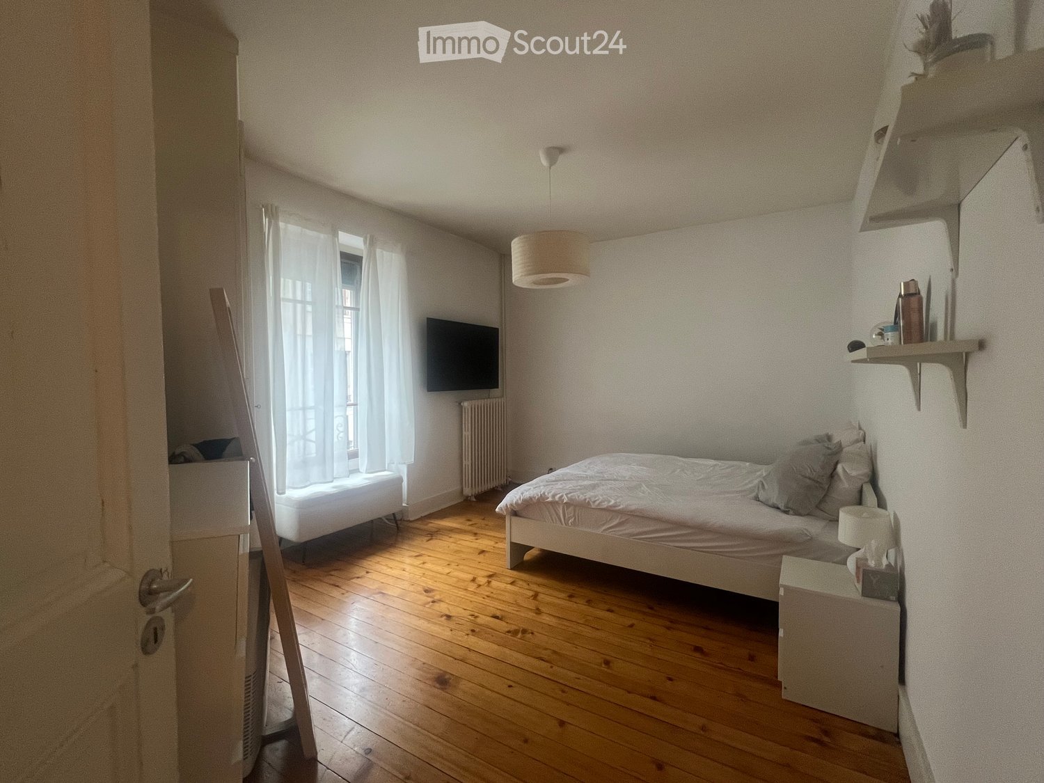 Cozy bedroom with wooden flooring, a bed with white bedding, a nightstand with a lamp, a television mounted on the wall, and a window with white curtains.