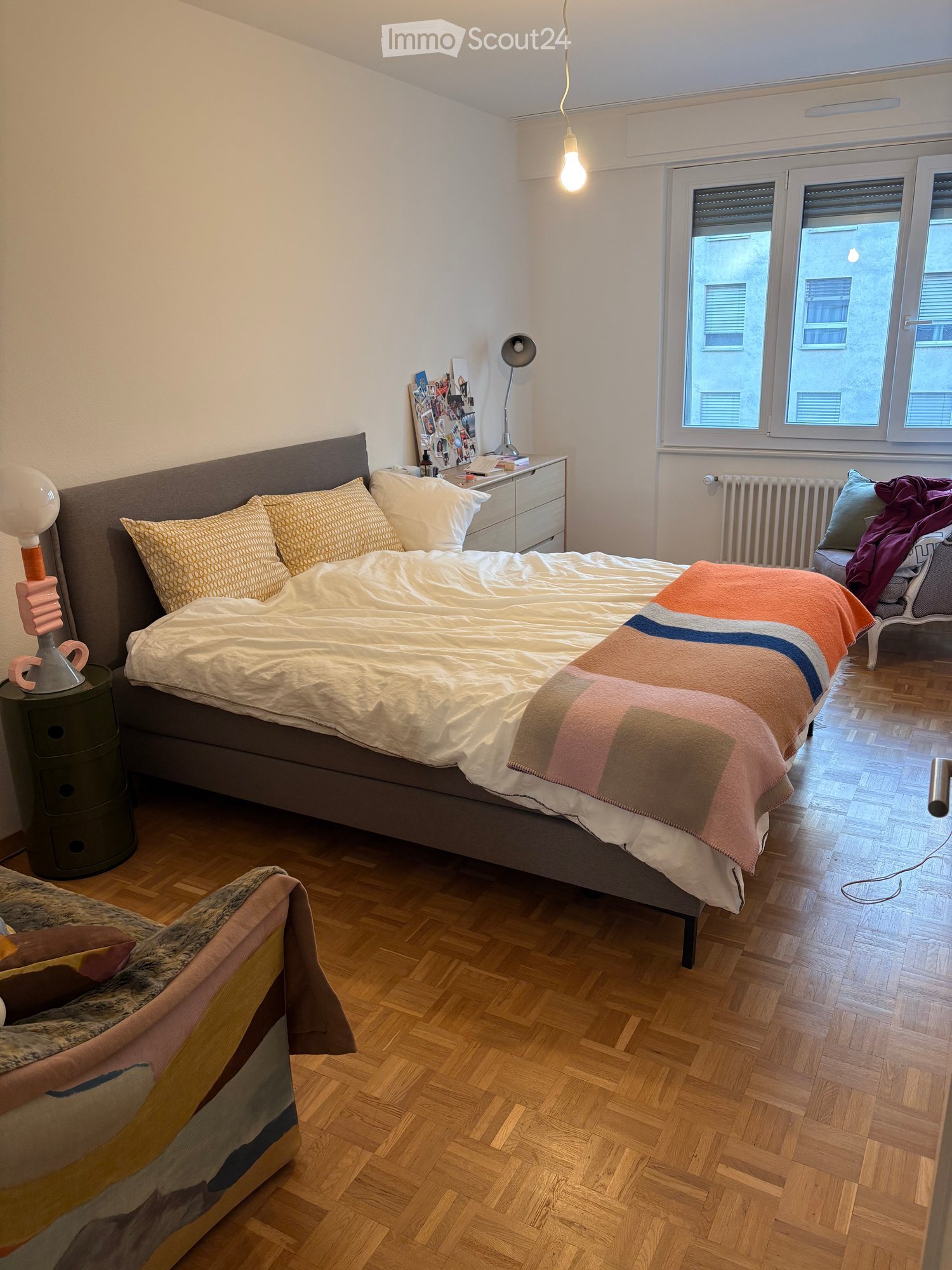 A modern bedroom with a large bed, colorful blanket, nightstand, lamp, and a chair with a blanket draped over it. The room features wooden flooring and large windows with blinds.