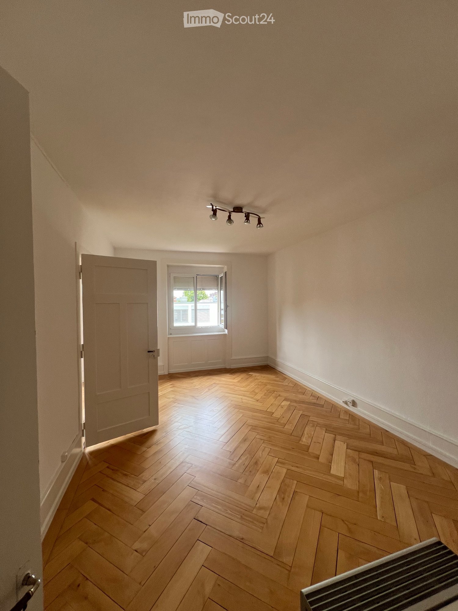 Empty room with wooden herringbone flooring, light-colored walls, a ceiling light fixture, a window with blinds, and a partially visible radiator.