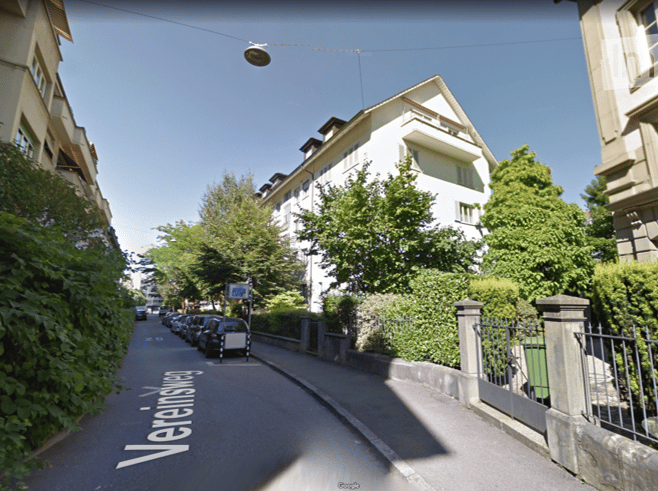 A quiet residential street lined with parked cars and multi-story apartment buildings, surrounded by lush green trees and shrubs.