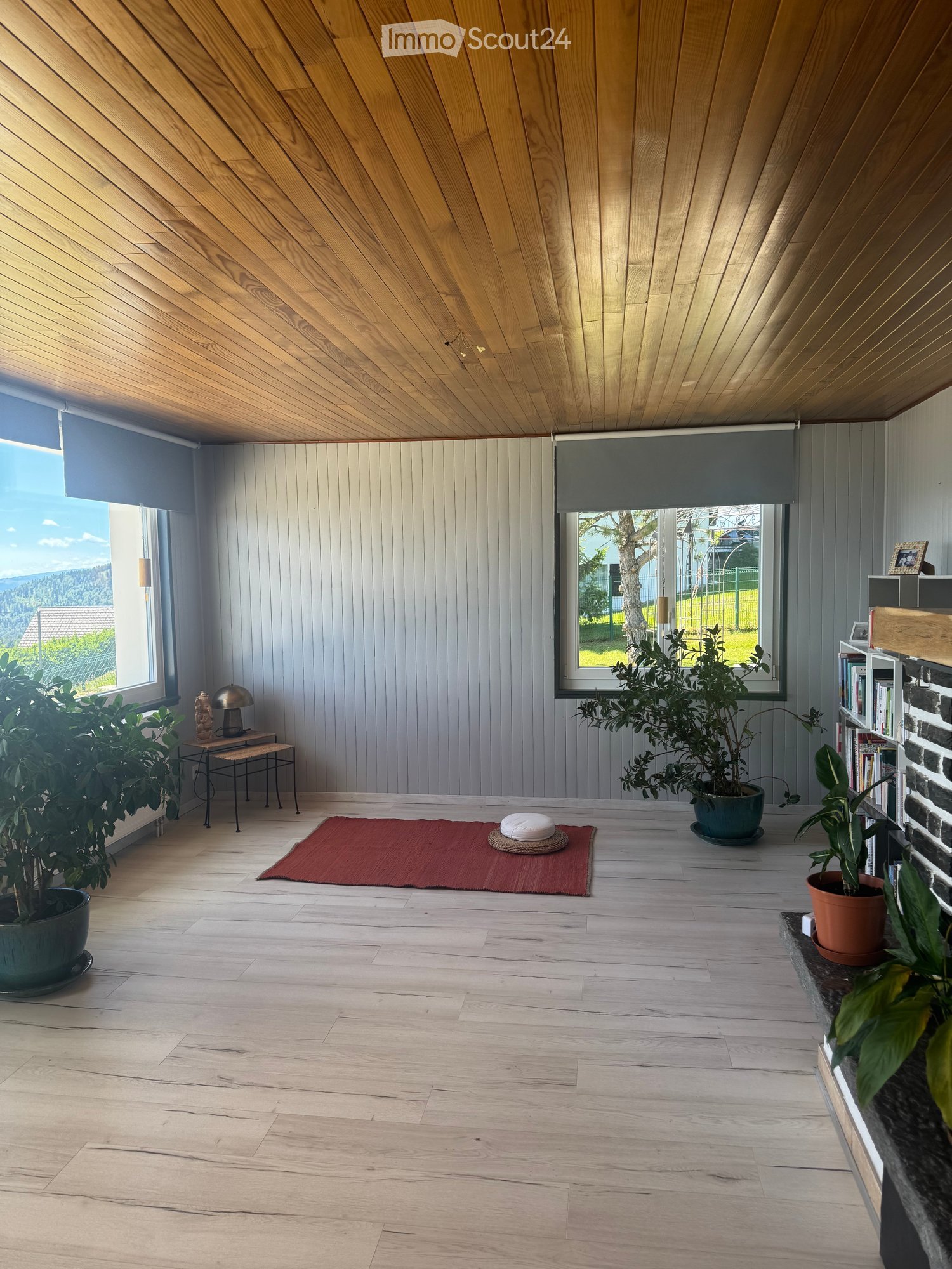 Bright, minimalist room with wooden floors and a slanted wooden ceiling. Features large windows with roller shades, a small table with a lamp, a bookshelf, potted plants, and a red meditation mat with cushions.