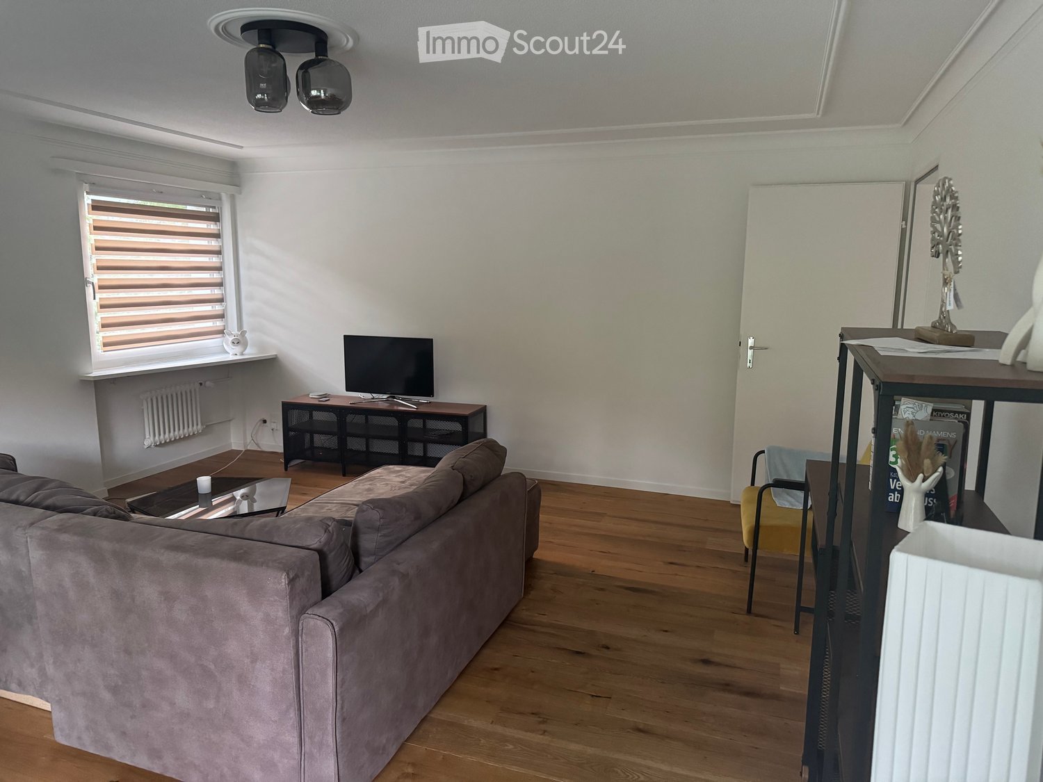 Modern living room with a grey sofa, coffee table, television, and a small table with a lamp. The room features wooden flooring and large windows with blinds.