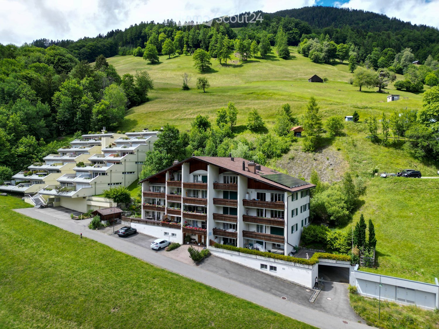 Aerial view of a multi-story residential building with balconies, surrounded by green fields and trees. The building has a modern design with a mix of white and brown facades, and several cars are parked in the driveway.