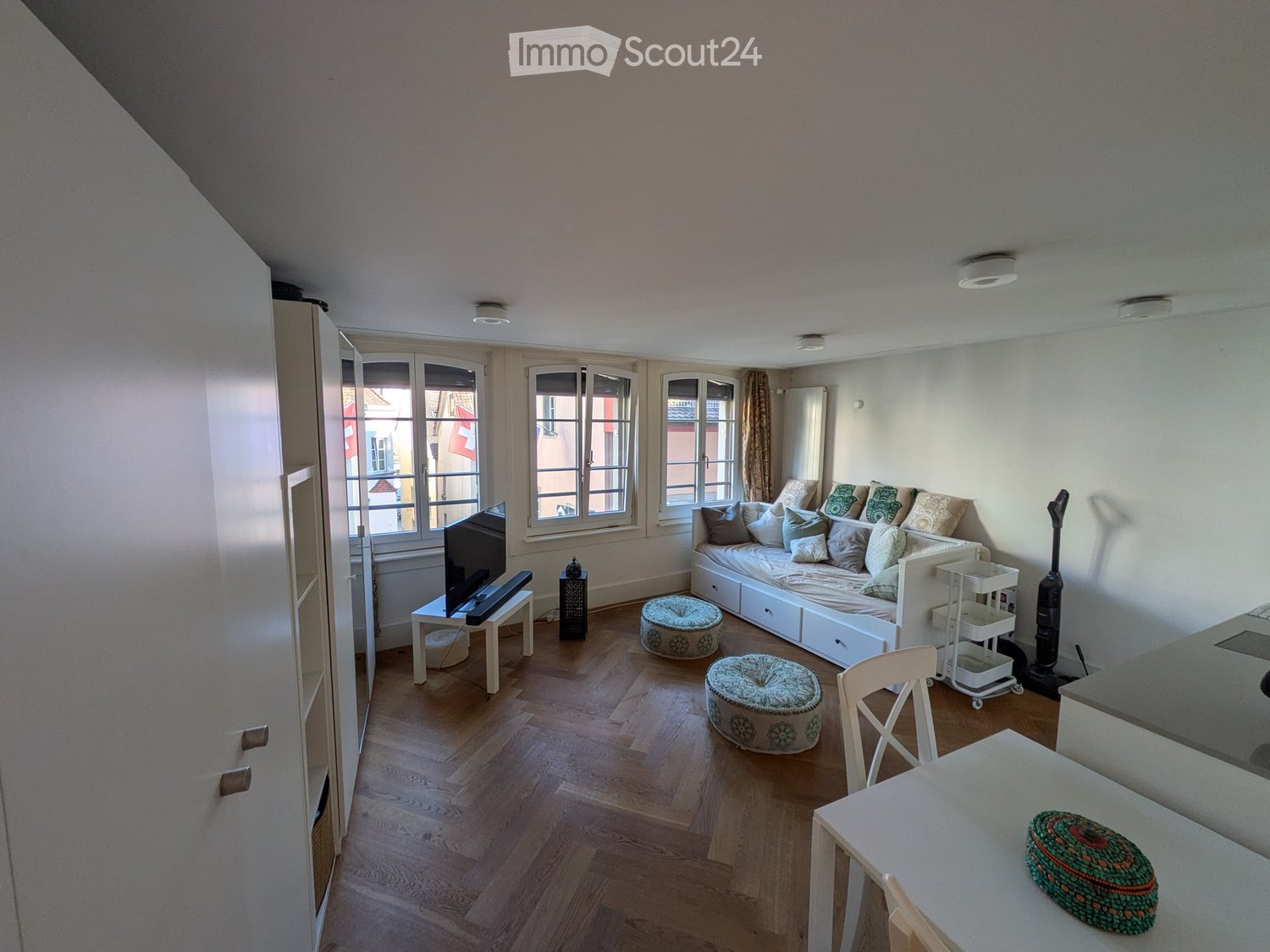 A modern, well-lit living space with wooden flooring, large windows, a sofa, a TV, and a small dining area.