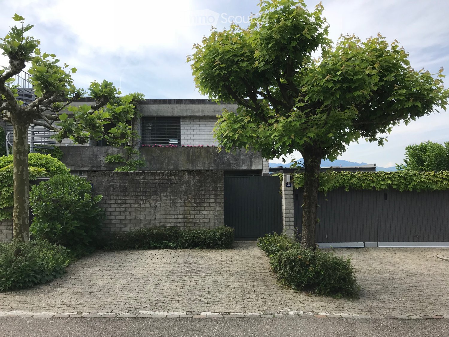 Modern two-story house with a concrete facade, large black garage door, and surrounding greenery including trees and shrubs.