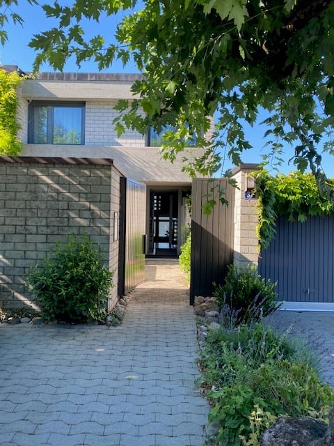 Modern two-story house with a clean, minimalist design. The entrance features a large, dark wooden door flanked by greenery and a paved walkway.