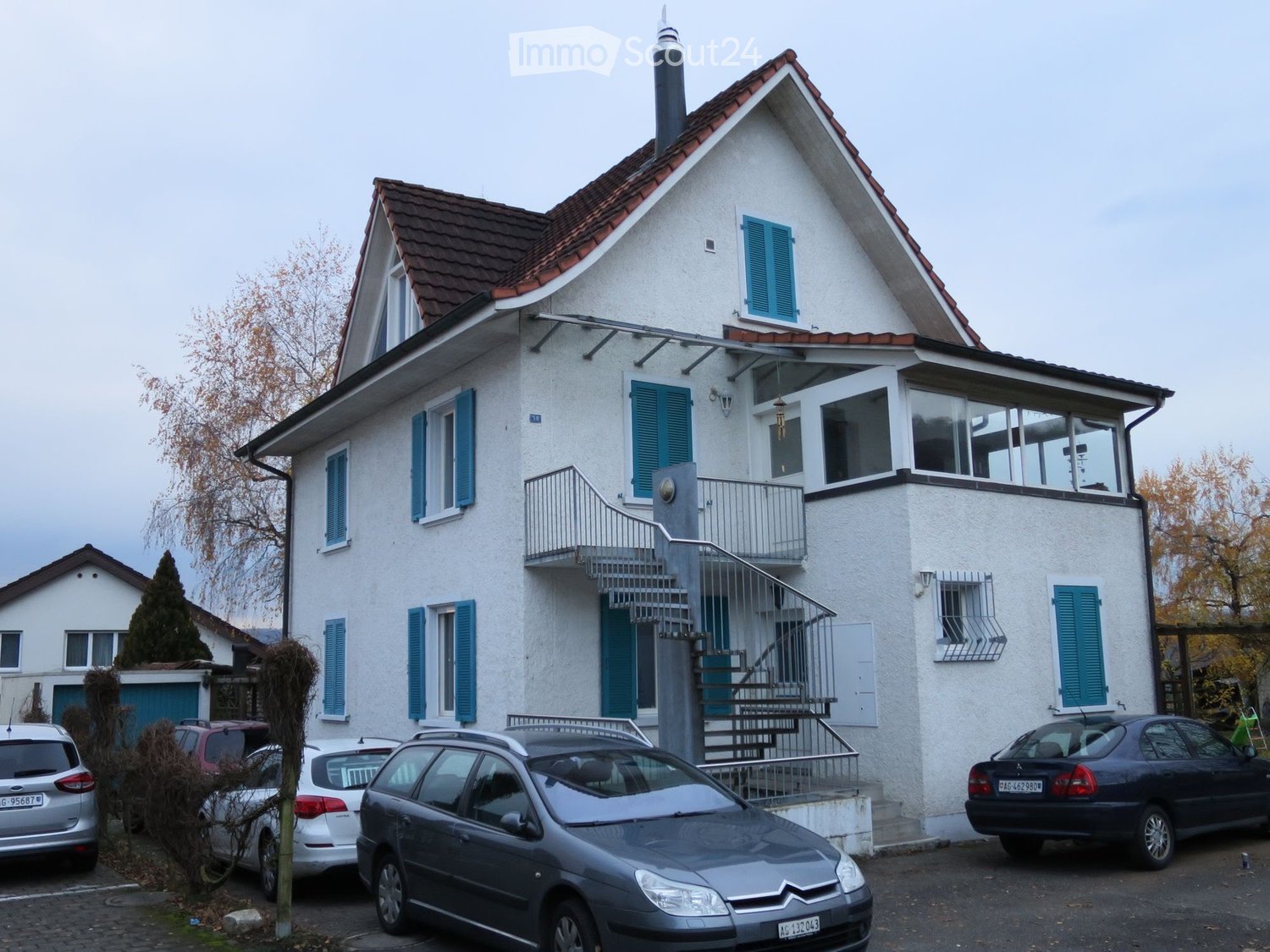 Two-story house with white exterior, blue shutters, and a balcony accessible via stairs. Several cars are parked in front.