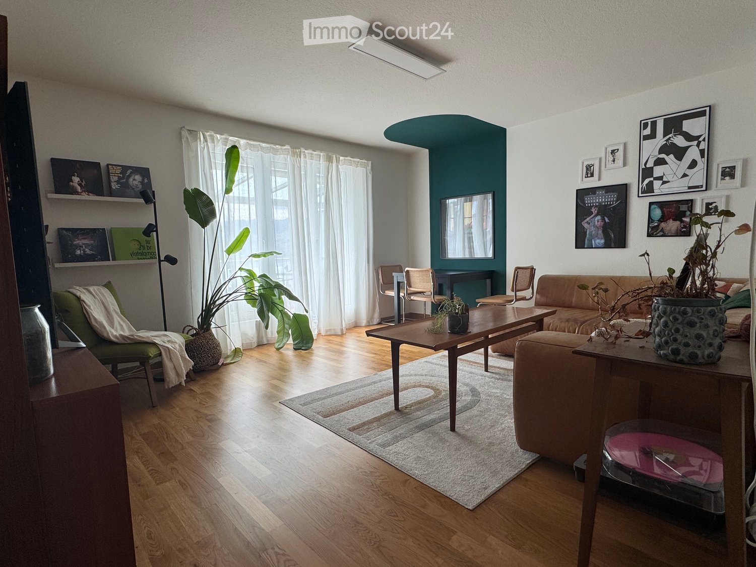 Modern living room with wooden flooring, a green accent wall, large windows with white curtains, a beige sofa, a wooden coffee table, a green armchair, and decorative shelves with artwork.