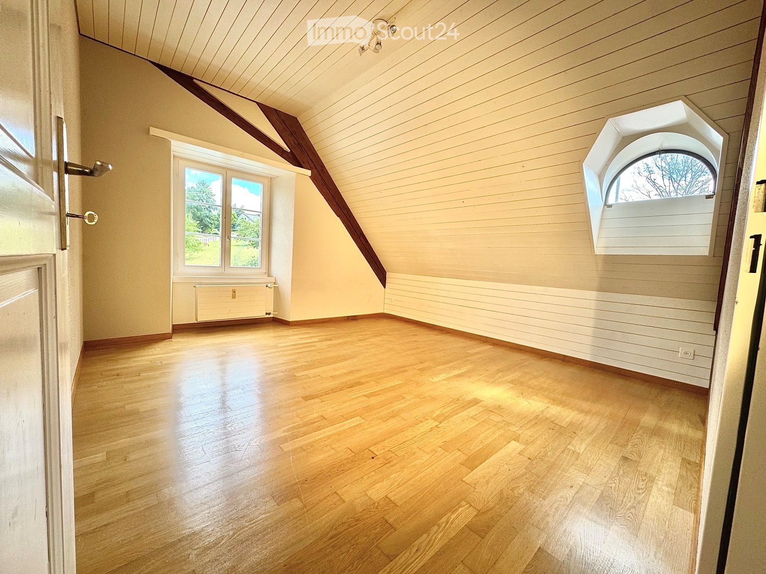 Empty room with wooden flooring, slanted ceiling, and large windows allowing natural light. The walls are painted in a light color, and there is a radiator under one window.