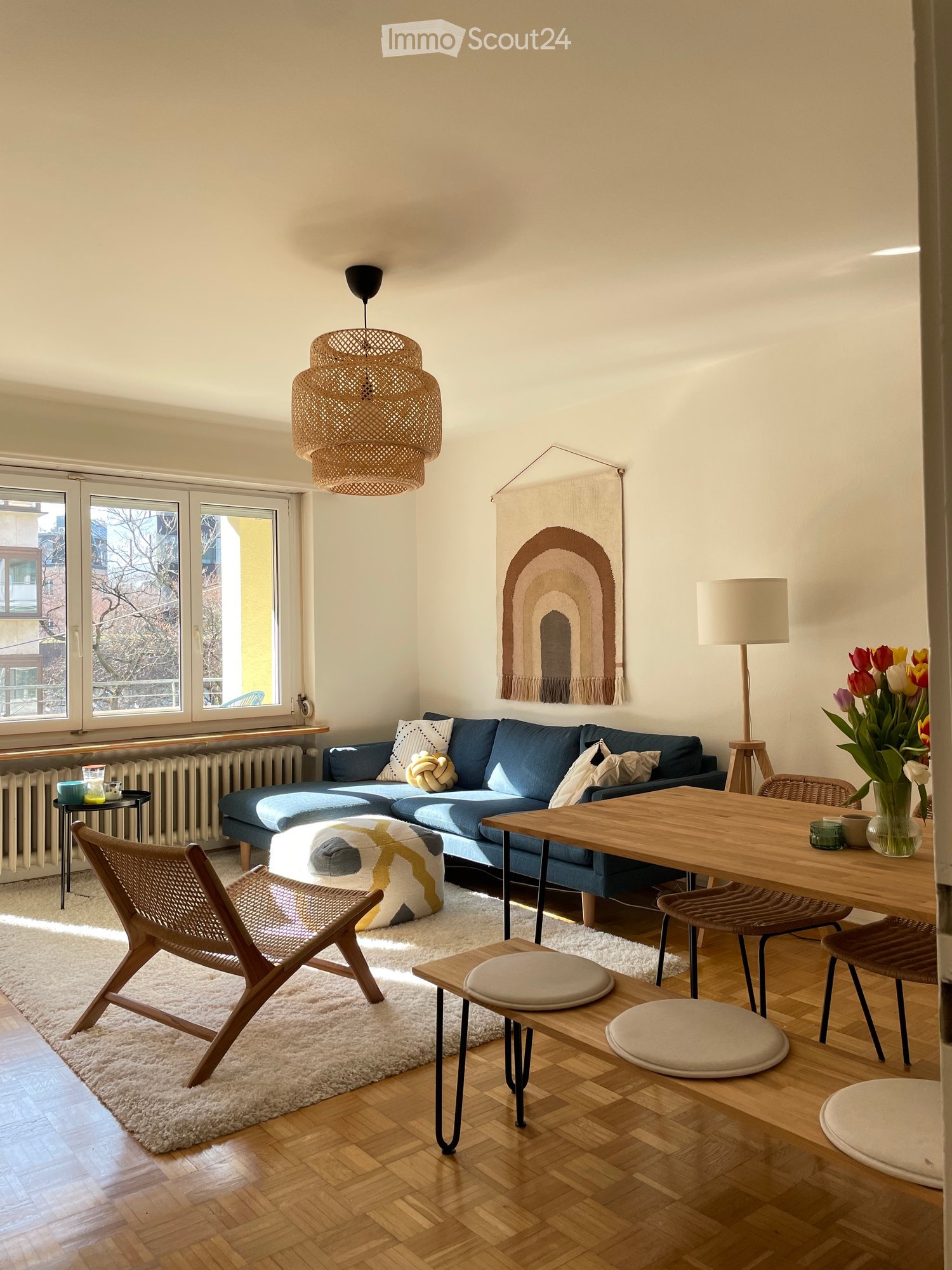 Modern living room with wooden flooring, white walls, and large windows. Features a blue sectional sofa, wooden dining table, woven pendant light, and decorative items.