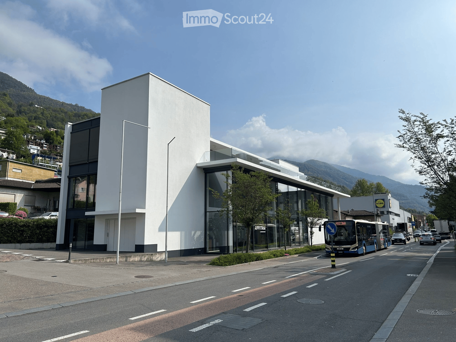 Modern white building with large glass windows and a sleek design, situated on a street with a bus passing by and a mountainous backdrop.