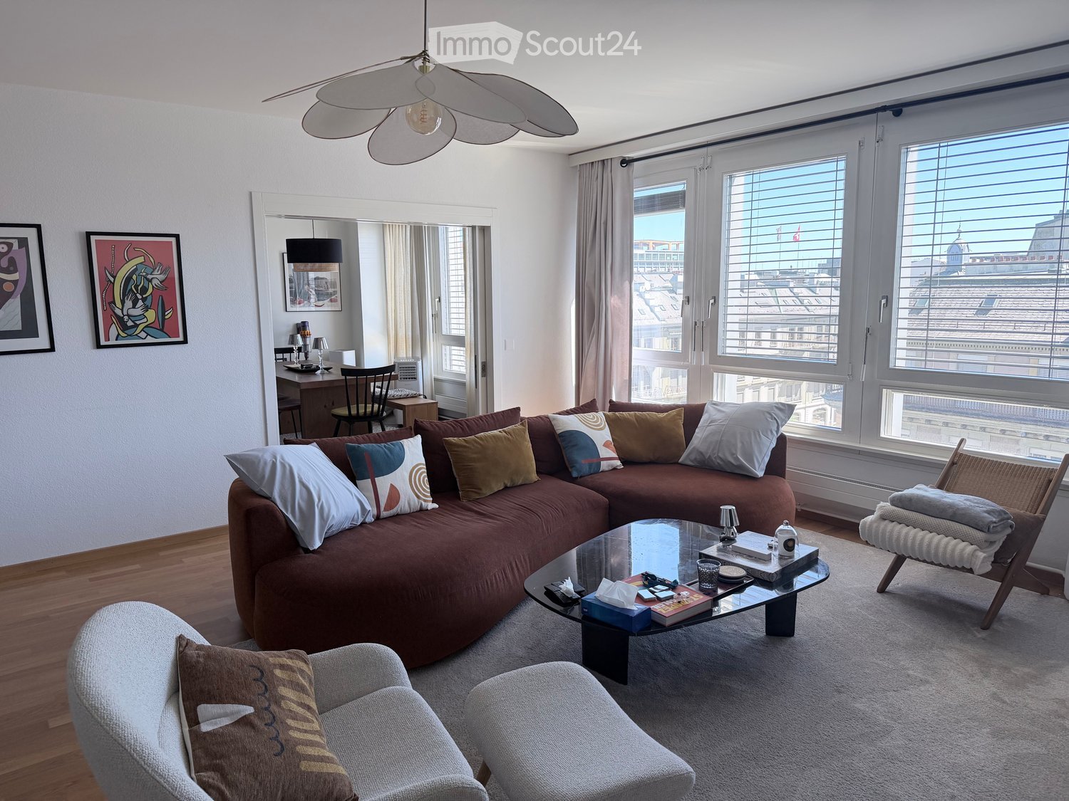Modern living room with large windows offering a city view, a brown sectional sofa with colorful pillows, a glass coffee table, a grey armchair, and a small desk area with a chair and lamp.