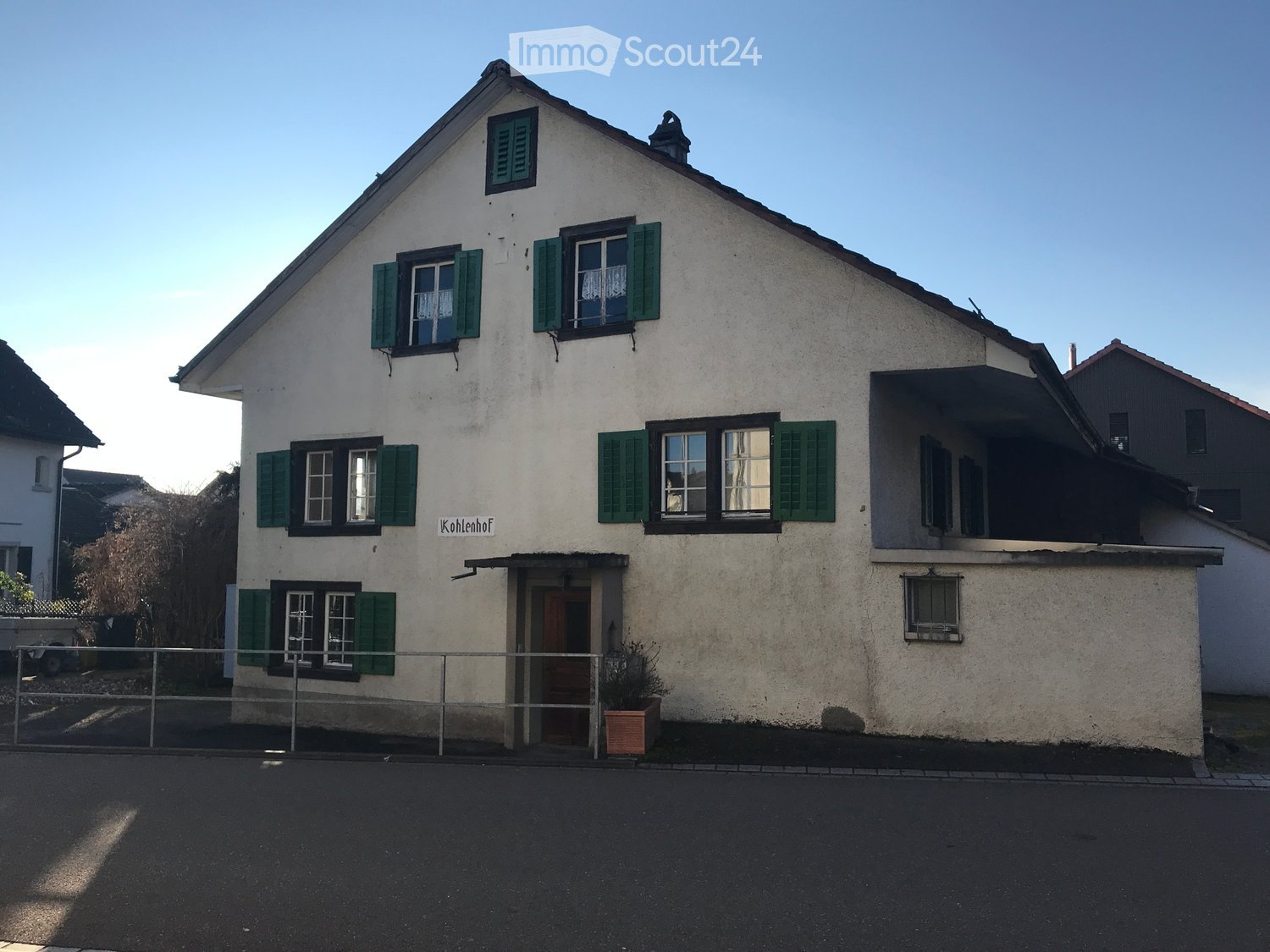 Two-story house with white plaster walls, green shutters, and a pitched roof. Features a covered entrance porch and a small balcony on the upper floor.
