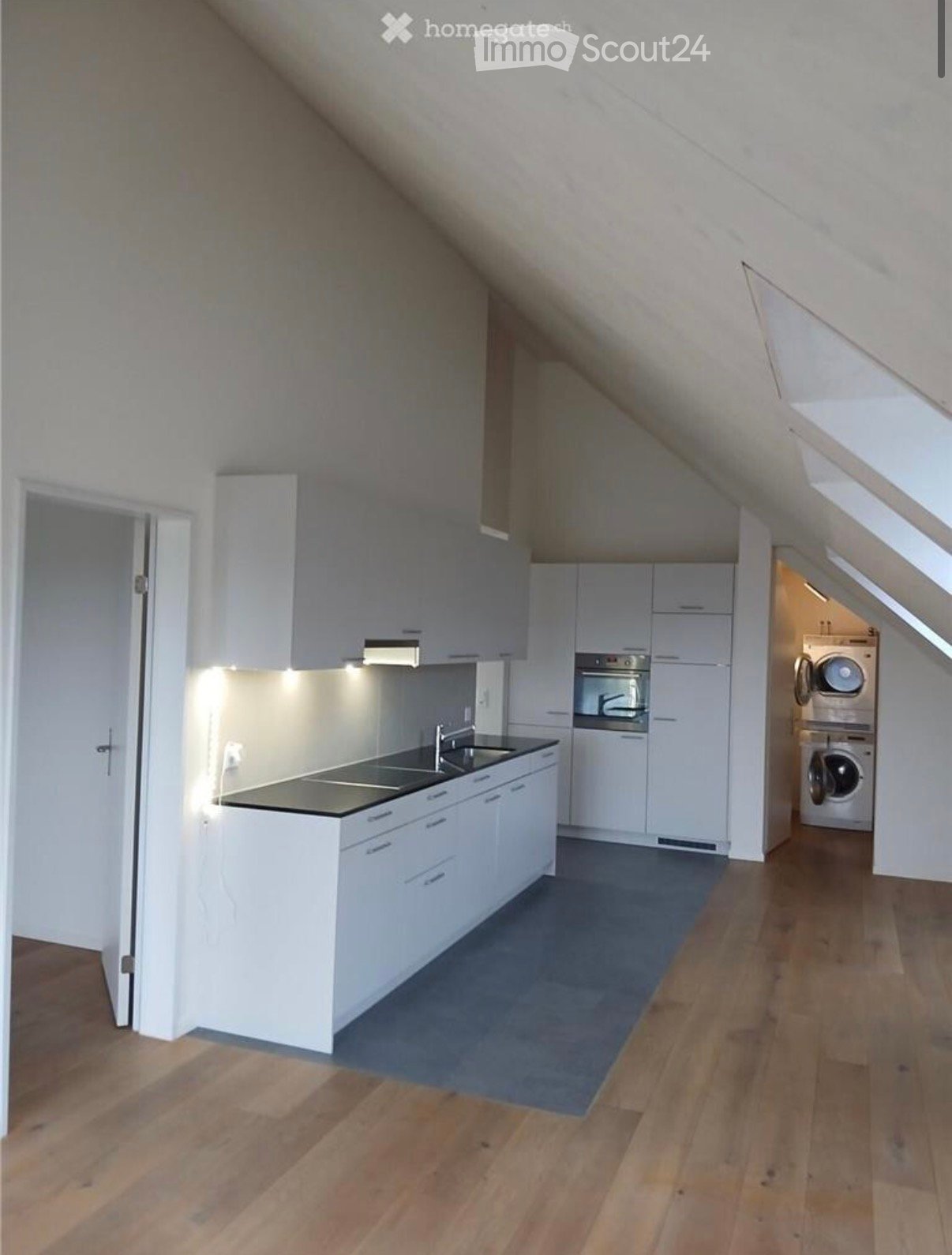 Modern kitchen with white cabinets, stainless steel sink, oven, and washing machine in an attic space with wooden flooring.