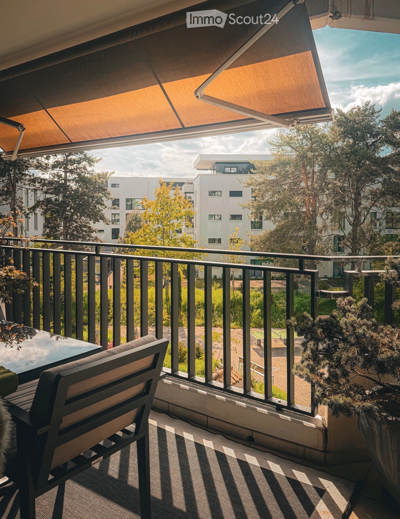 Balcony with a bench, table, and potted plants, featuring a retractable awning and a view of a modern residential area.