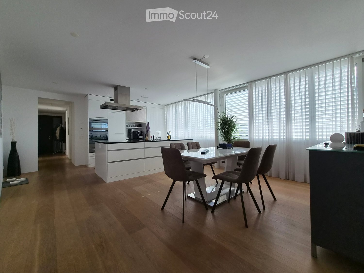 Modern kitchen and dining area with white cabinets, a central island, a dining table with chairs, and large windows with blinds.