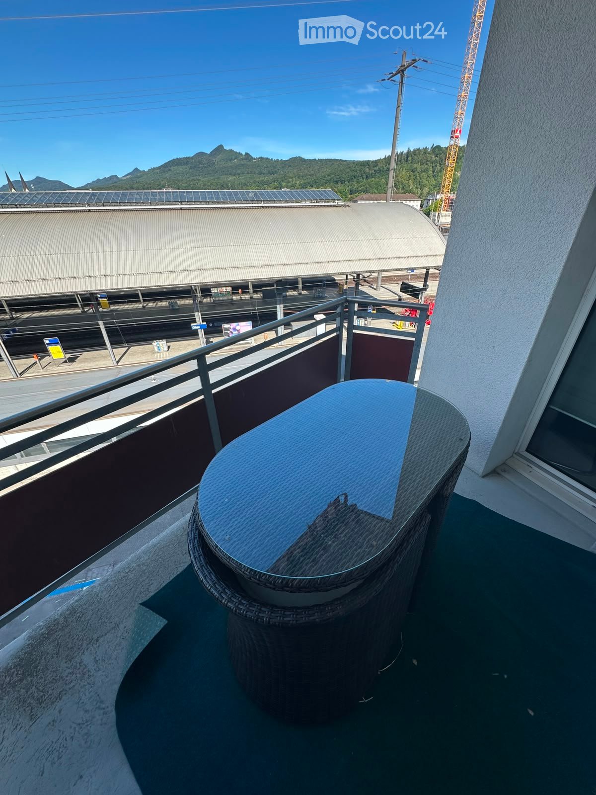 Balcony with a round wicker table and a metal railing, overlooking a train station and distant mountains.