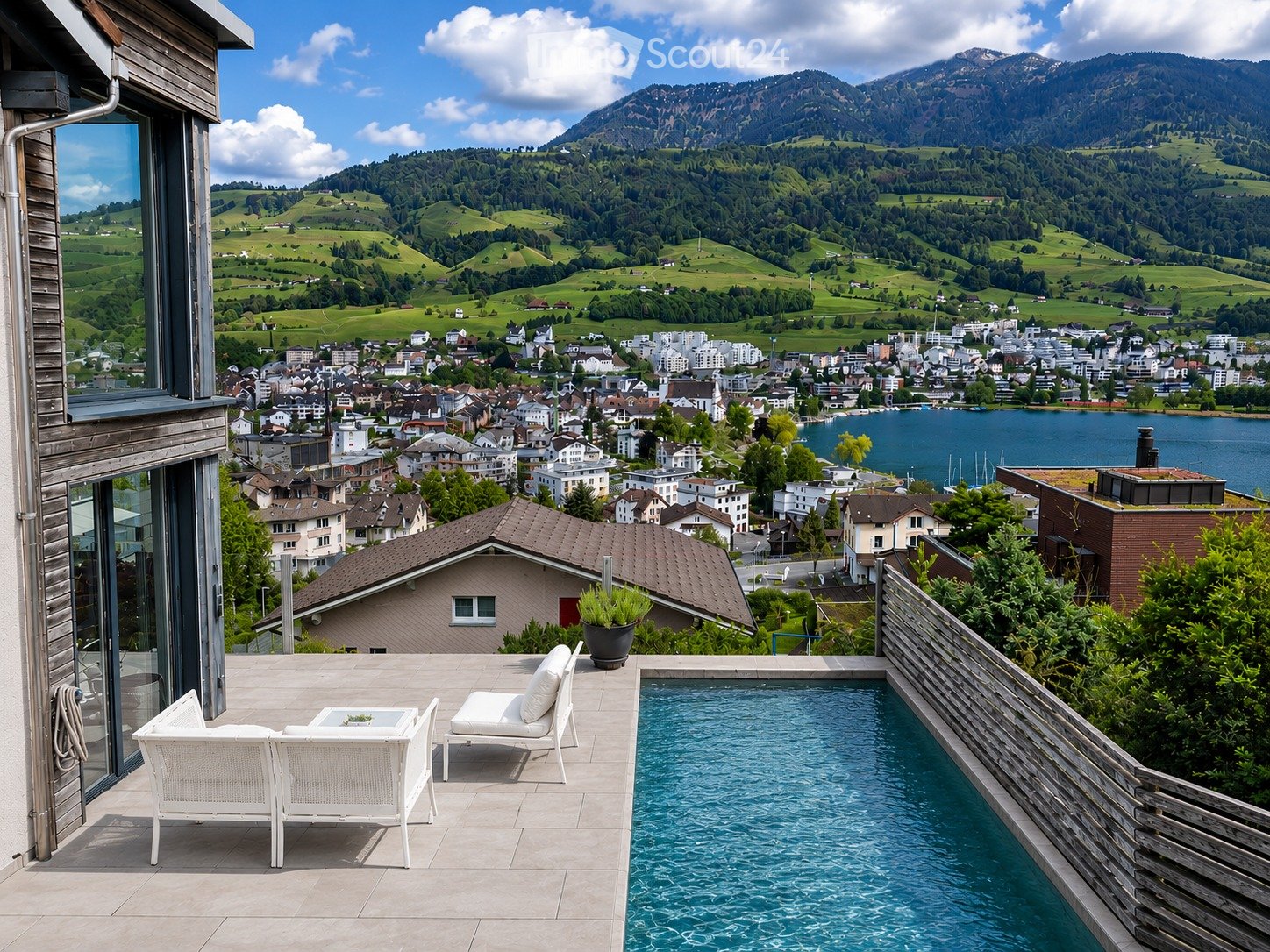Balcony with a swimming pool, white lounge chairs, a small table, and a scenic view of a town and mountains.